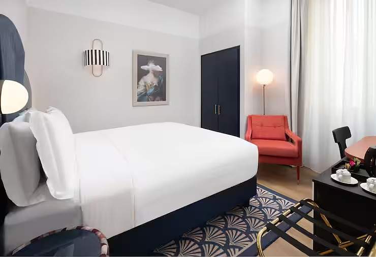Bed dressed in white against a navy base, below a framed portrait and a striped wall light, with a coral armchair and a fan-pattern rug