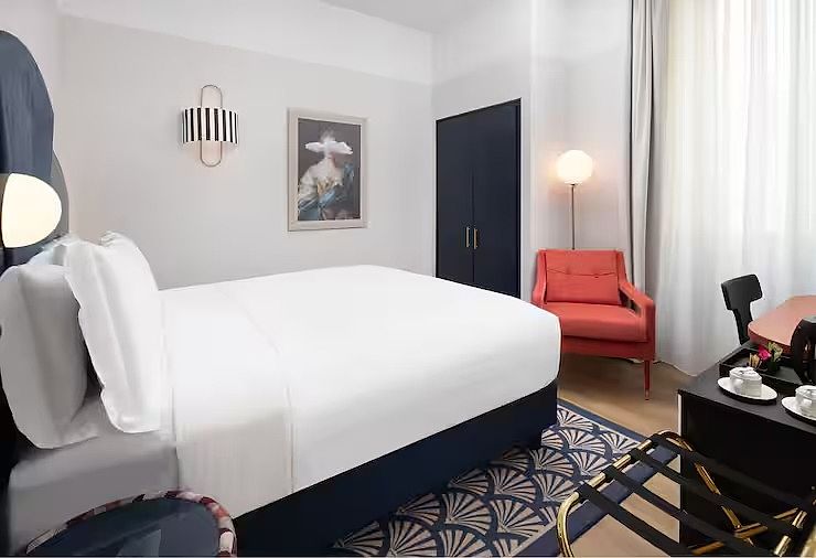 Bed dressed in white against a navy base, below a framed portrait and a striped wall light, with a coral armchair and a fan-pattern rug
