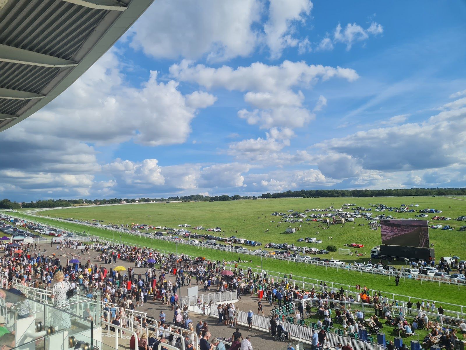 Crowds on the Hill at Epsom Downs seen from a grandstand balcony, with parked cars, a big screen and the Downs stretching away behind