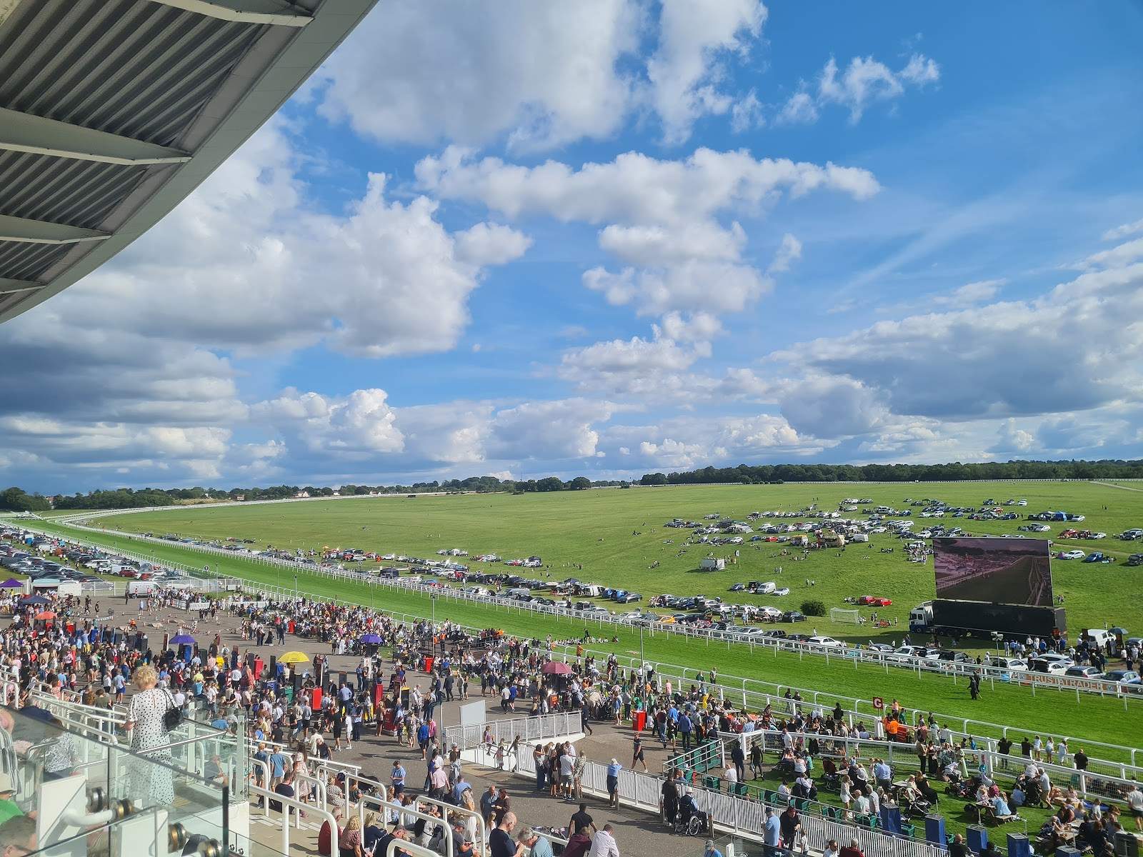 Crowds on the Hill at Epsom Downs seen from a grandstand balcony, with parked cars, a big screen and the Downs stretching away behind