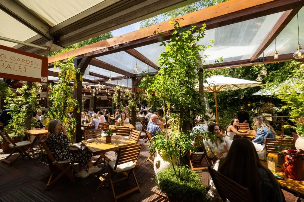Guests at wooden tables under a glass roof and climbing plants in the Spring Garden at the Chelsea Flower Show, beside a sign for the garden chalet