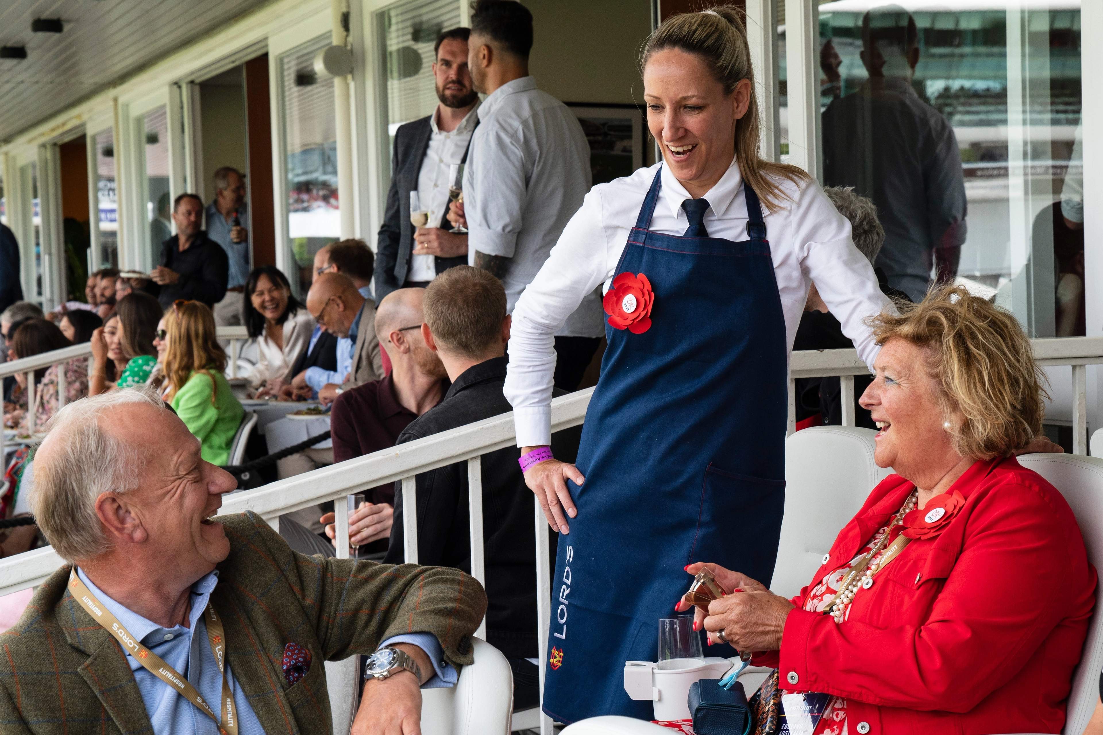 Guests in a hospitality box at Lord's, one in a green tweed check jacket and open-necked shirt, another in red, with a host in a Lord's apron leaning in to talk to them