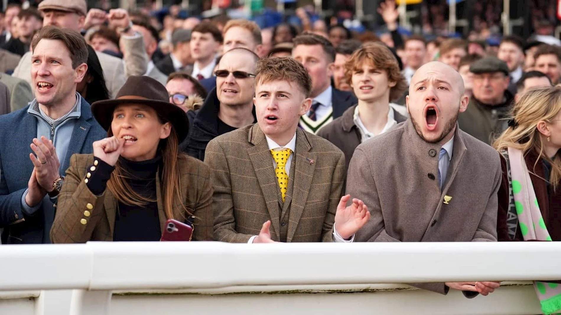 Racegoers at the rail at Cheltenham in the country dress code, including a woman in a felt hat and tweed jacket and a man in a checked tweed suit