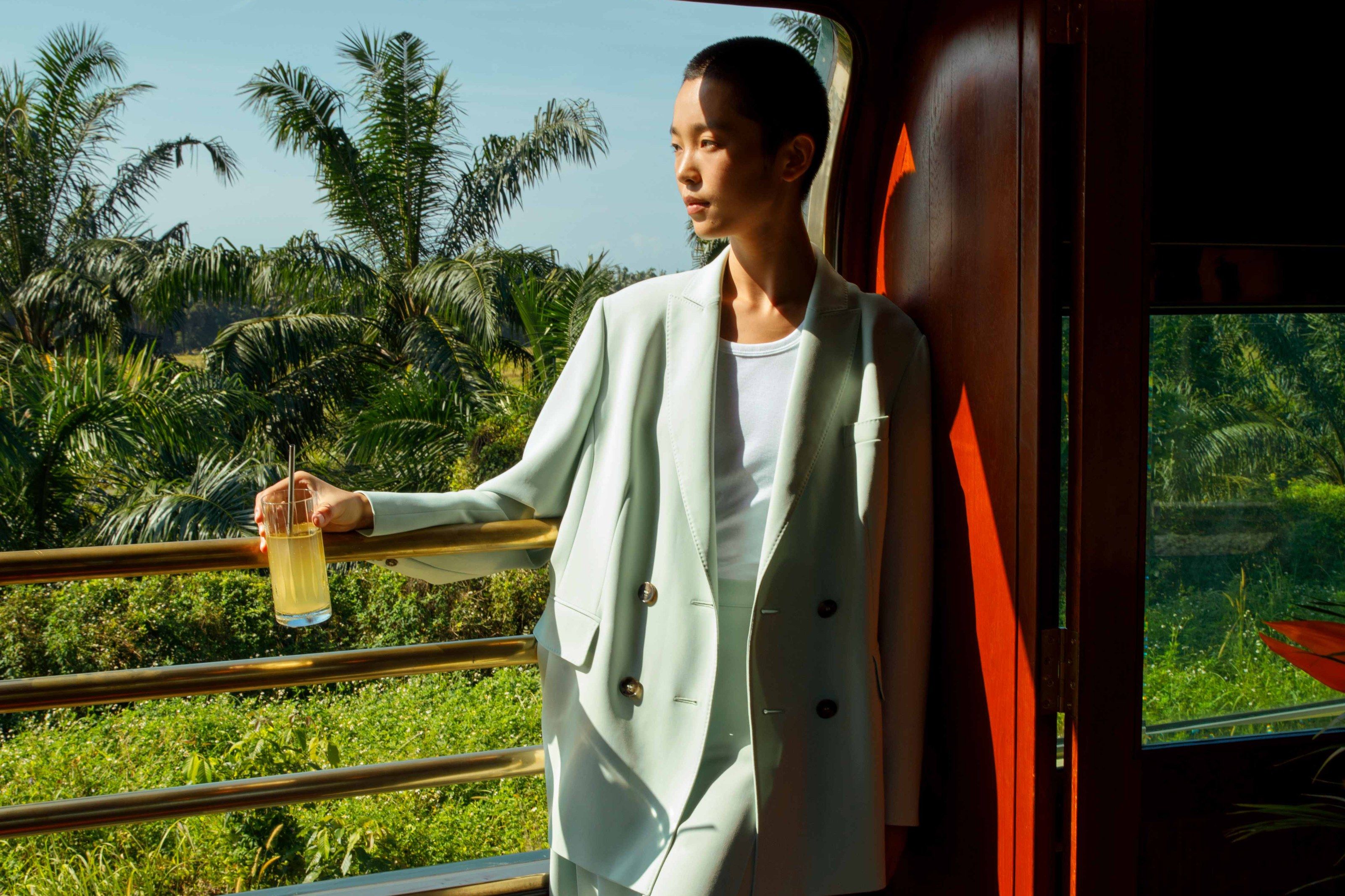 A guest holding a drink at the open side of the observation car, with palms and green fields behind.