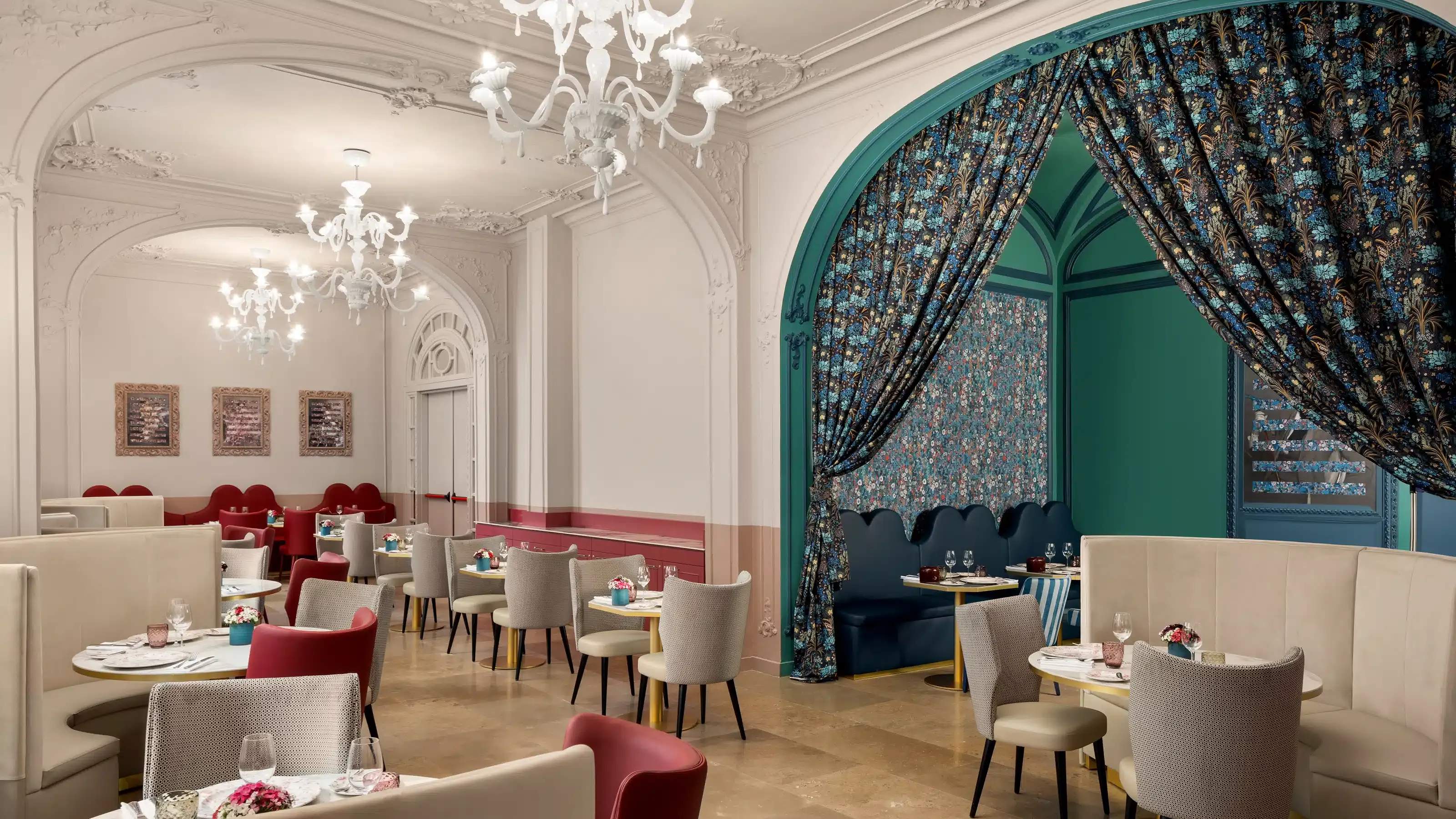 Restaurant dining room with white glass chandeliers, cream banquettes and red chairs at round tables, and a teal archway hung with floral curtains over a booth