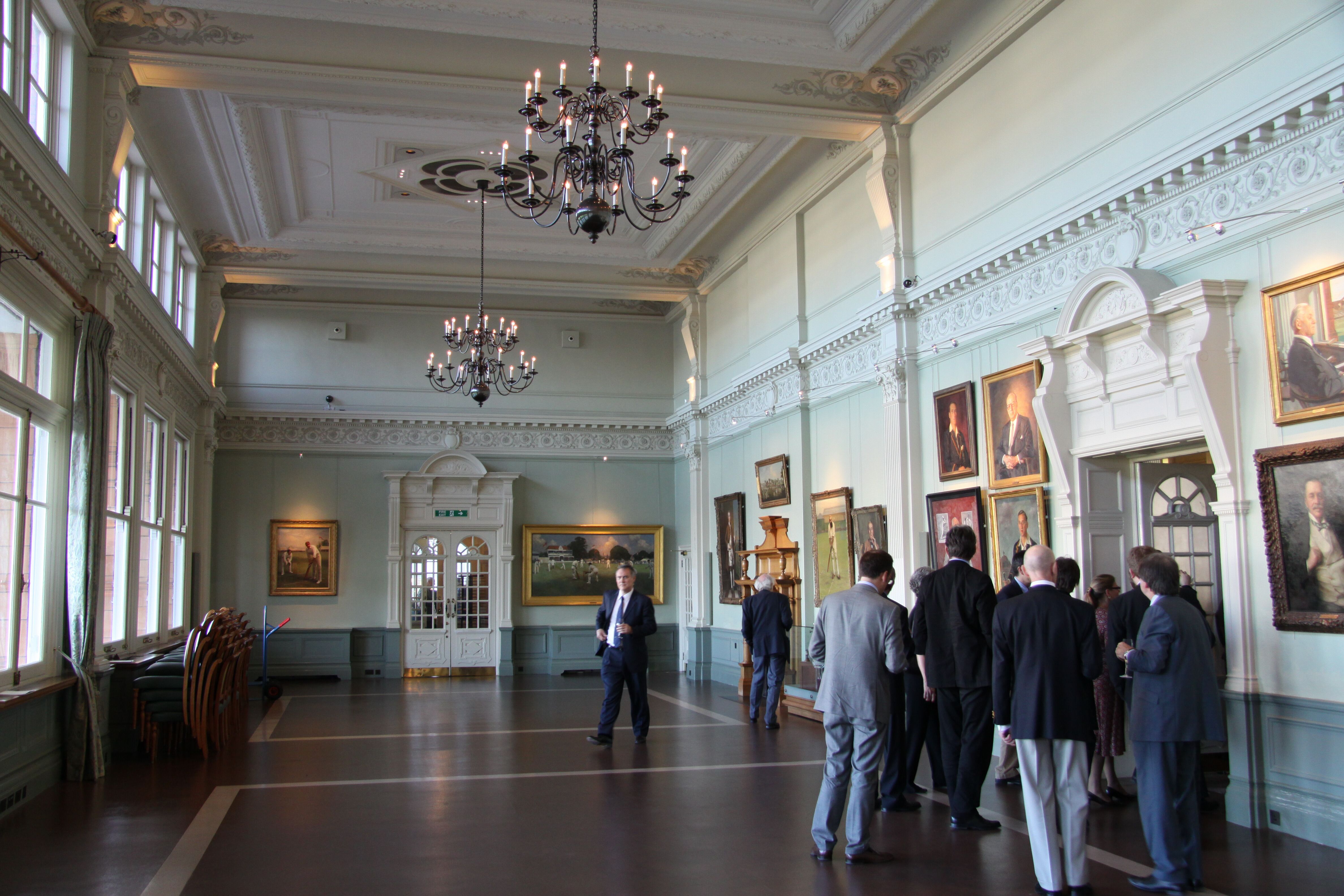 The Long Room at Lord's, a long panelled hall hung with paintings, with a group of men in suits and jackets standing at the far end