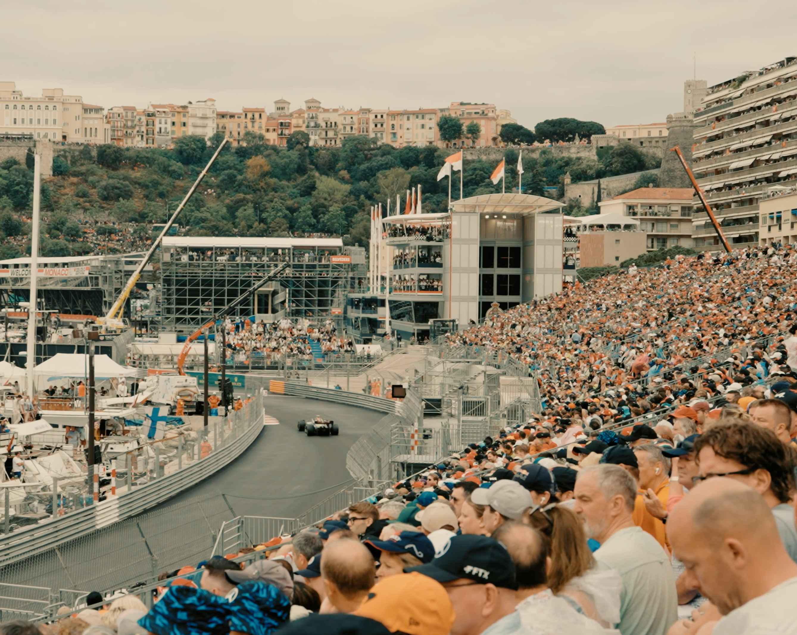 Spectators in a grandstand on Port Hercule watching a Formula 1 car pass along the harbour, with moored boats and the Rock of Monaco behind