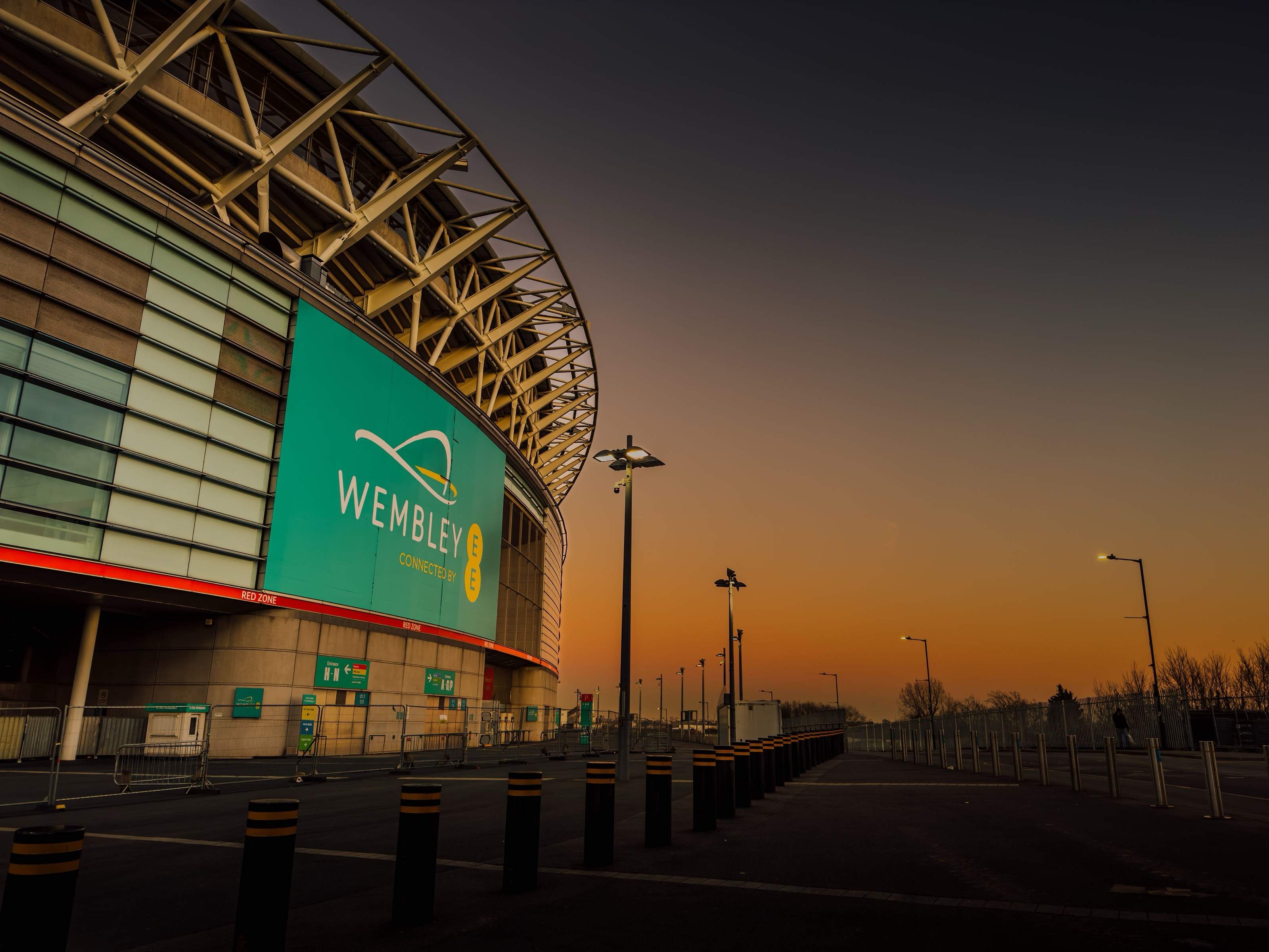 Wembley Stadium's curved front and Wembley sign at sunset, under an orange sky