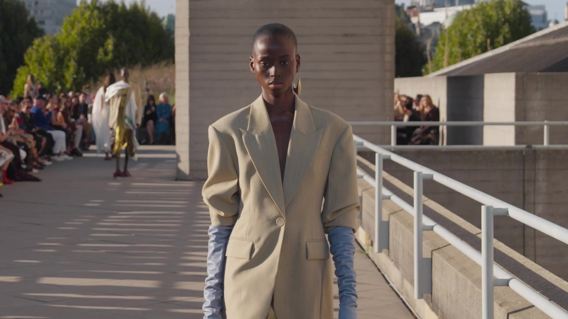 A model in camel tailoring and pale blue gloves on the runway at Roksanda's September 2026 show, guests seated behind