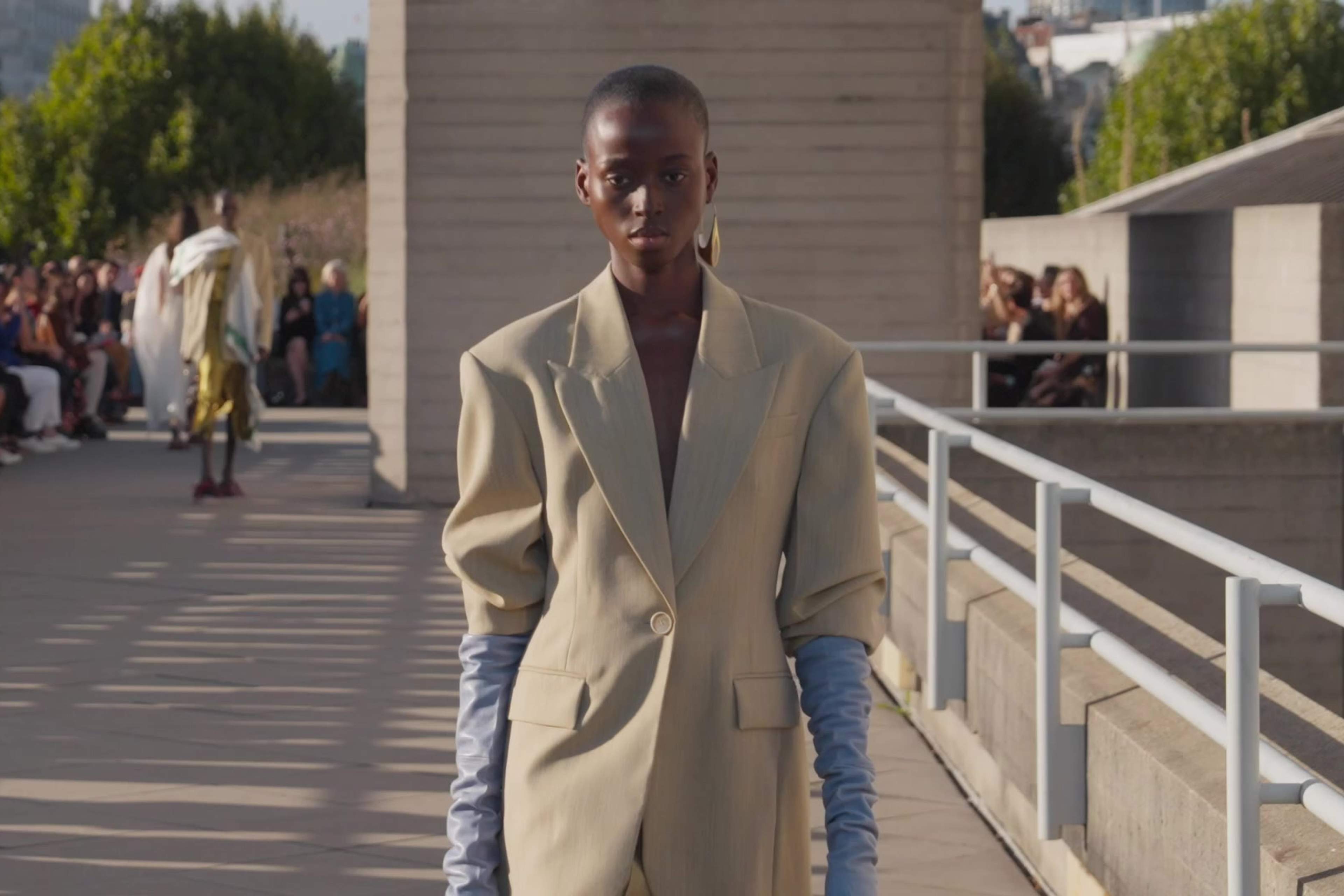 A model in camel tailoring and pale blue gloves on the runway at Roksanda's September 2026 show, guests seated behind