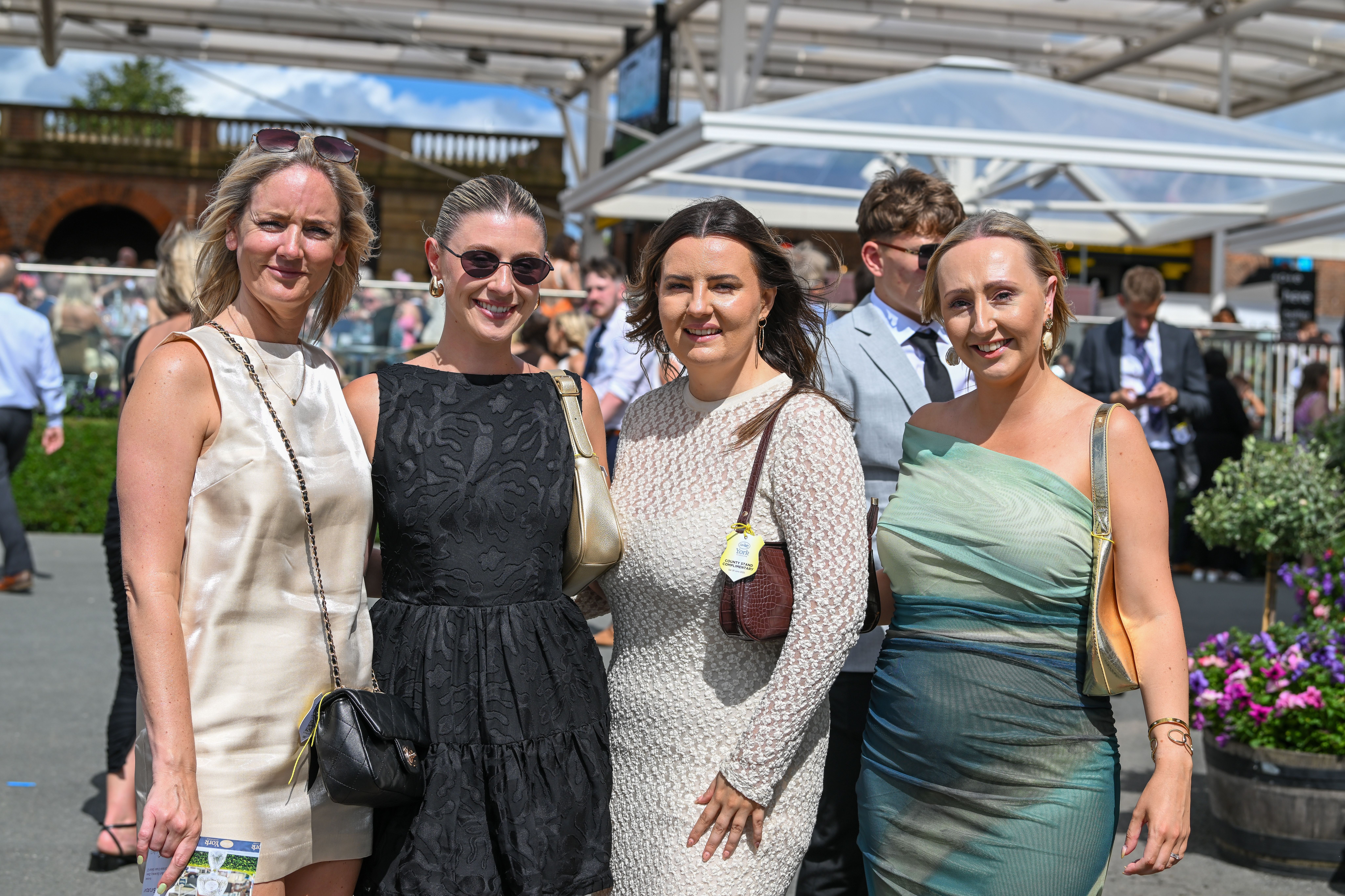 Four guests in summer dresses standing together on a sunny raceday at York Racecourse, one holding a racecard