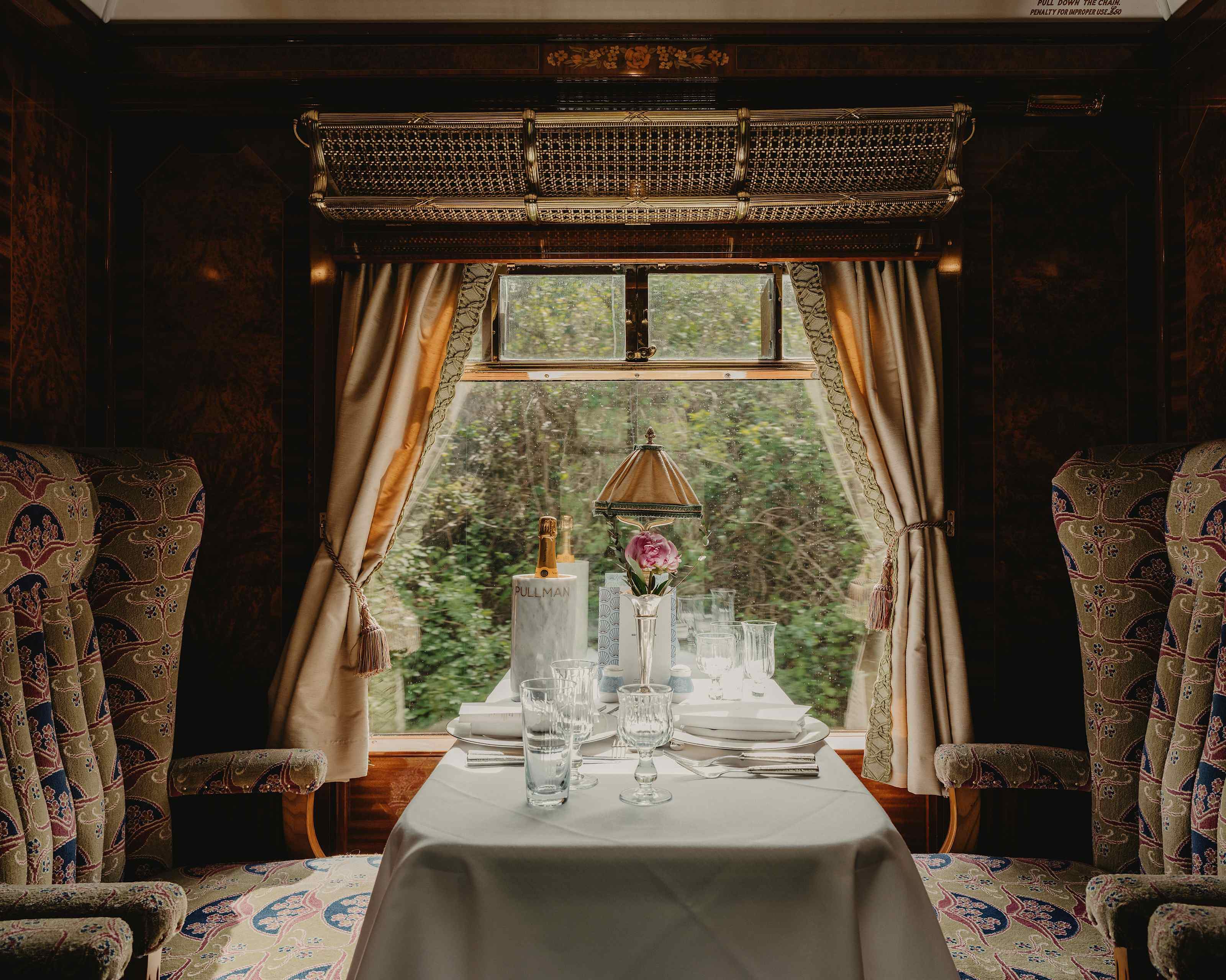 A table for two laid by a carriage window, with a lamp and flowers
