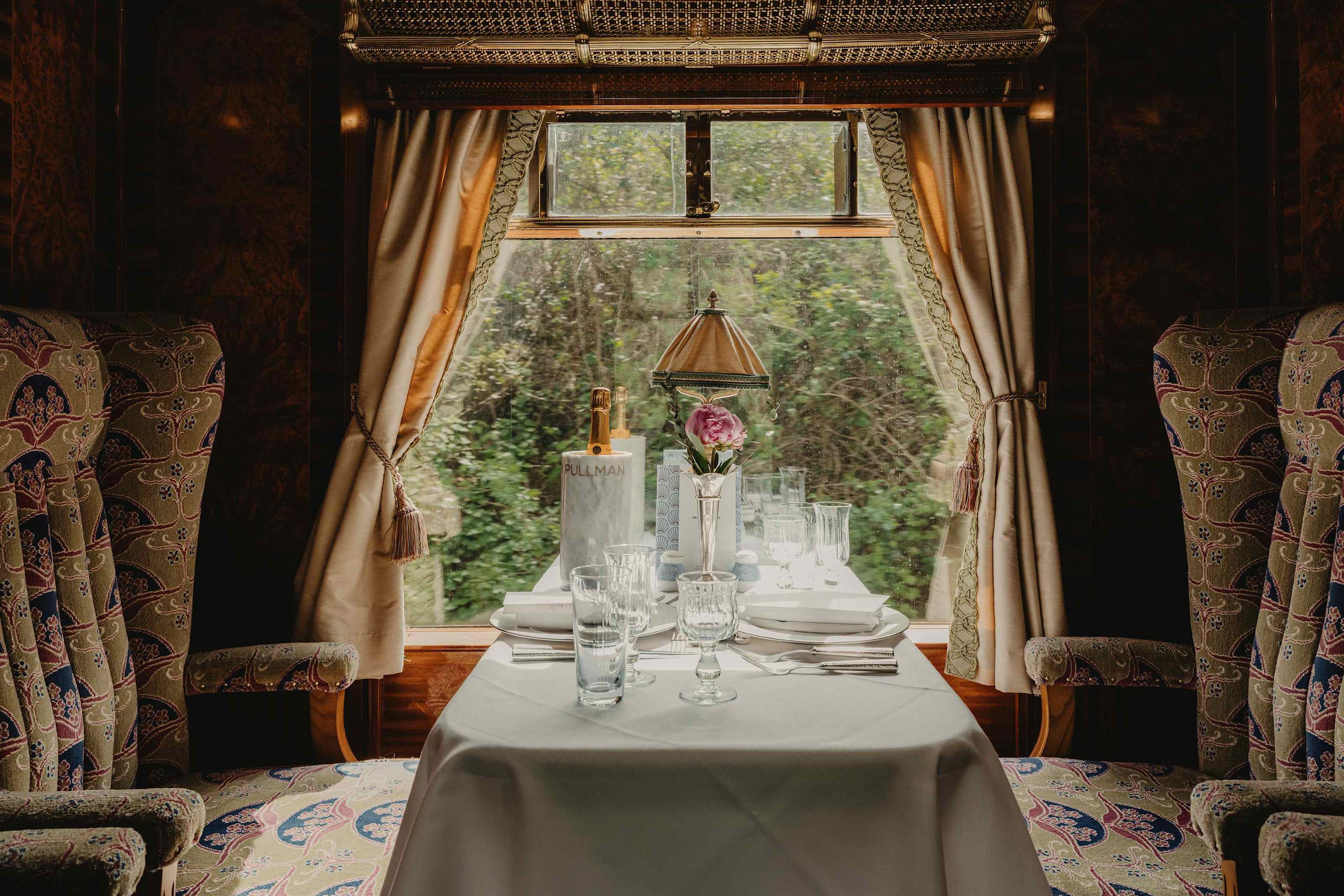 A table for two laid by a carriage window, with a lamp and flowers