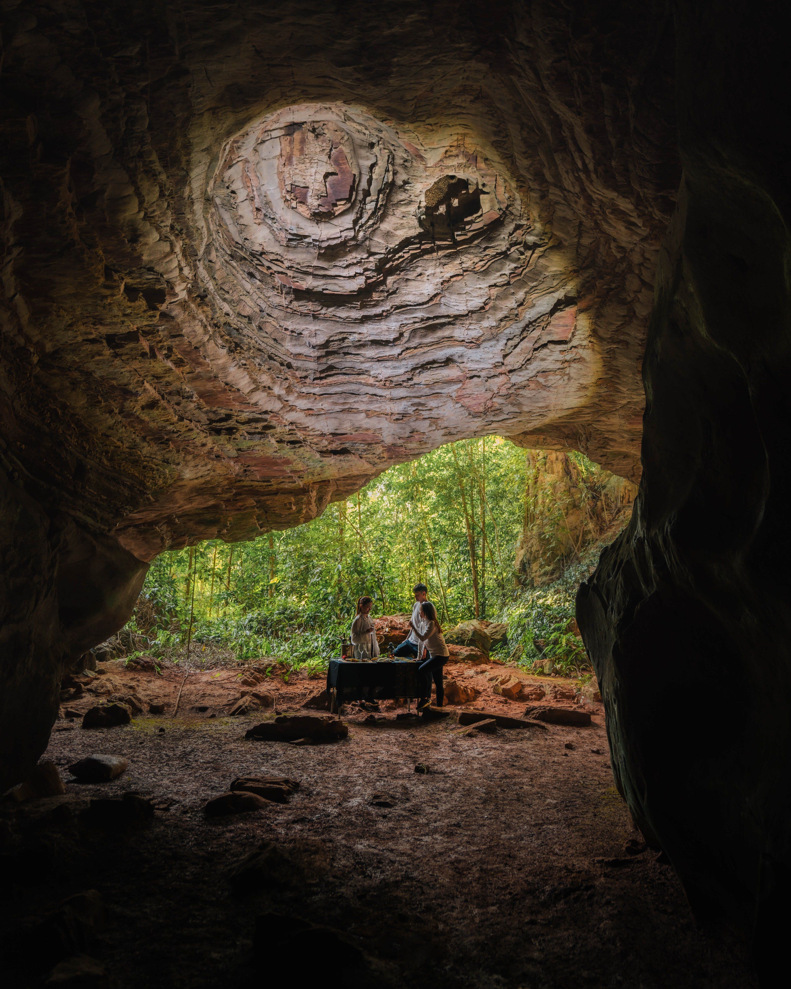 Two guests at a table inside the mouth of a limestone cave near Merapoh, with forest beyond.