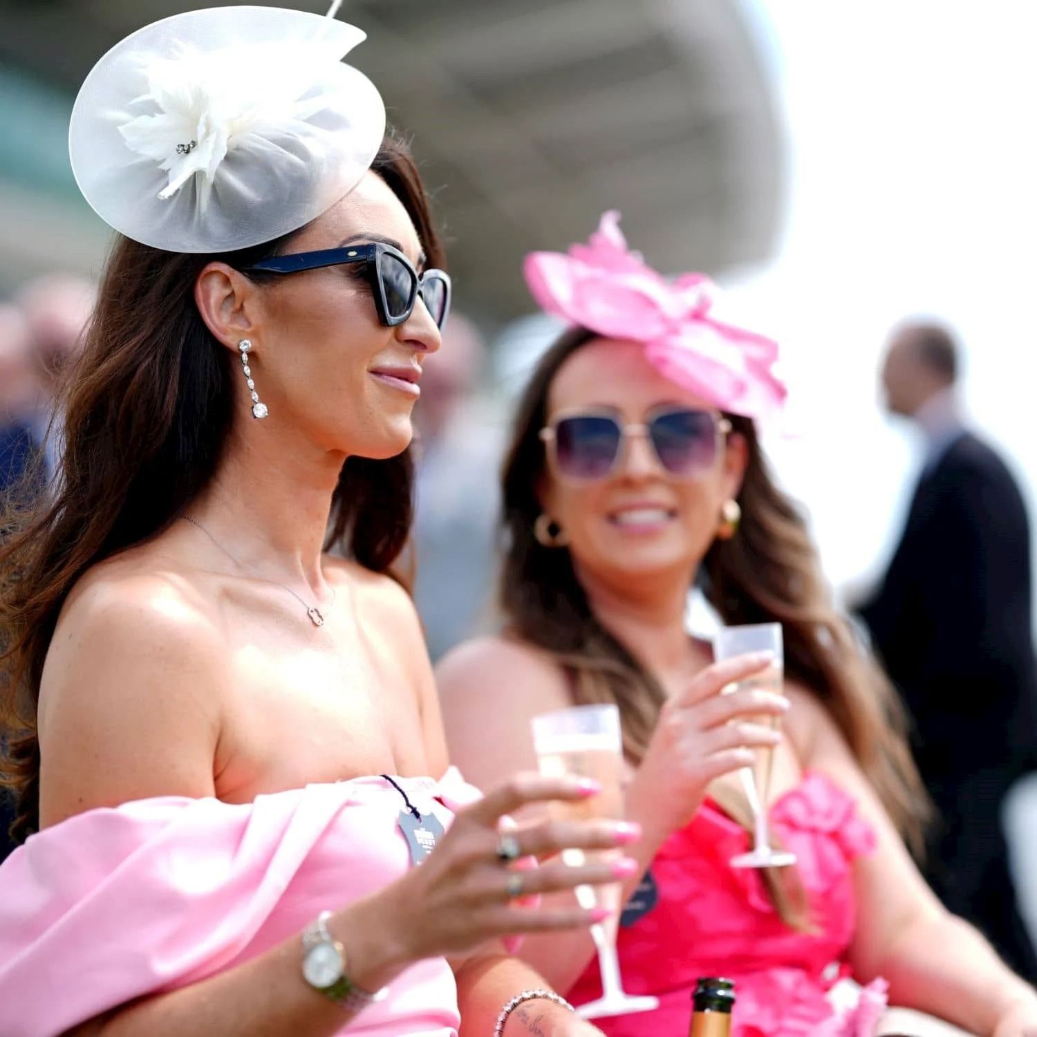 Two women at Epsom in summer dresses and fascinators, one white and one pink, holding glasses of champagne, with a grandstand behind them