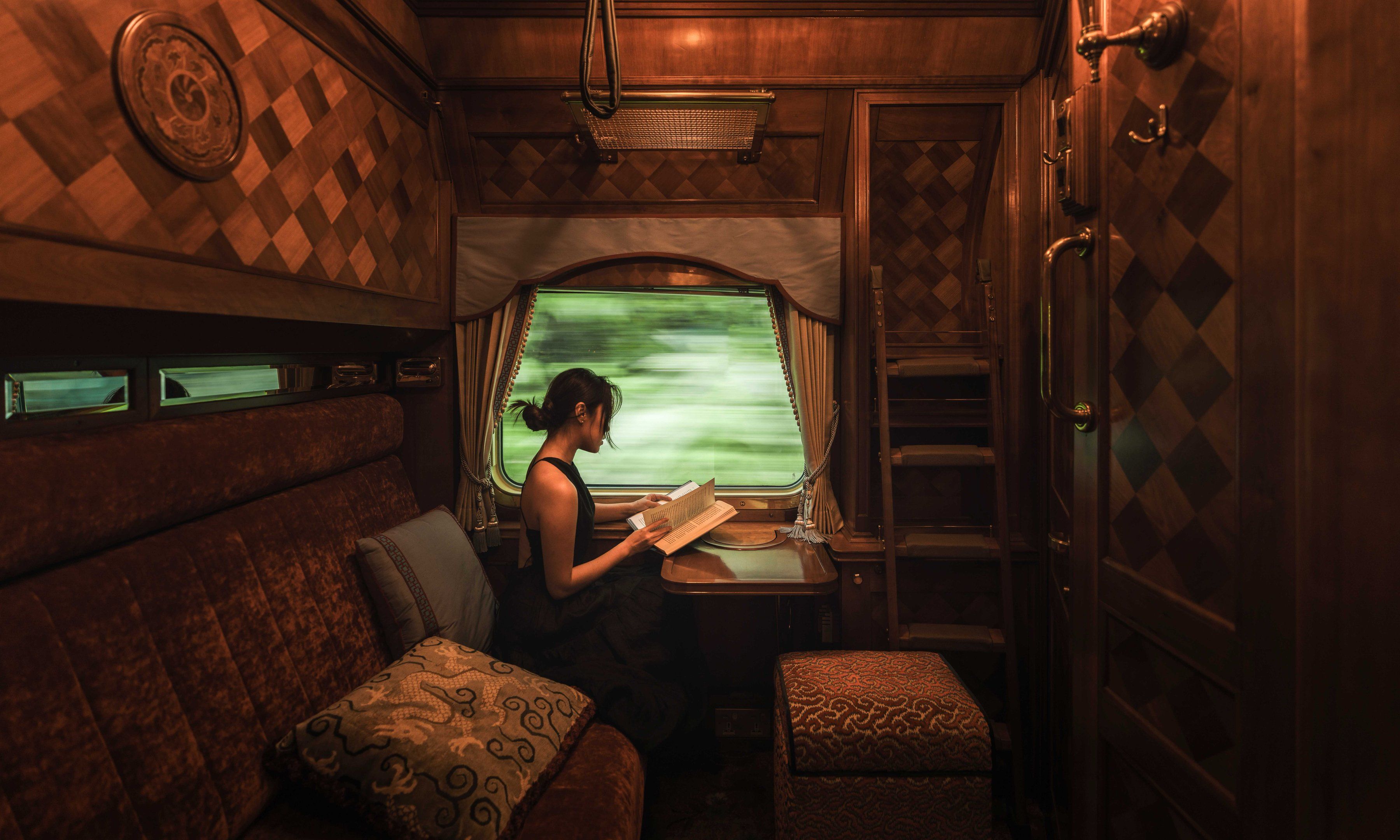 A guest reads a book at the window of a wood-panelled Pullman Cabin on the Eastern & Oriental Express, with green forest blurring past outside.