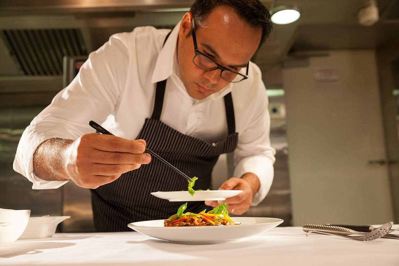 A chef in a white shirt and dark apron plating a dish with tweezers in a restaurant kitchen