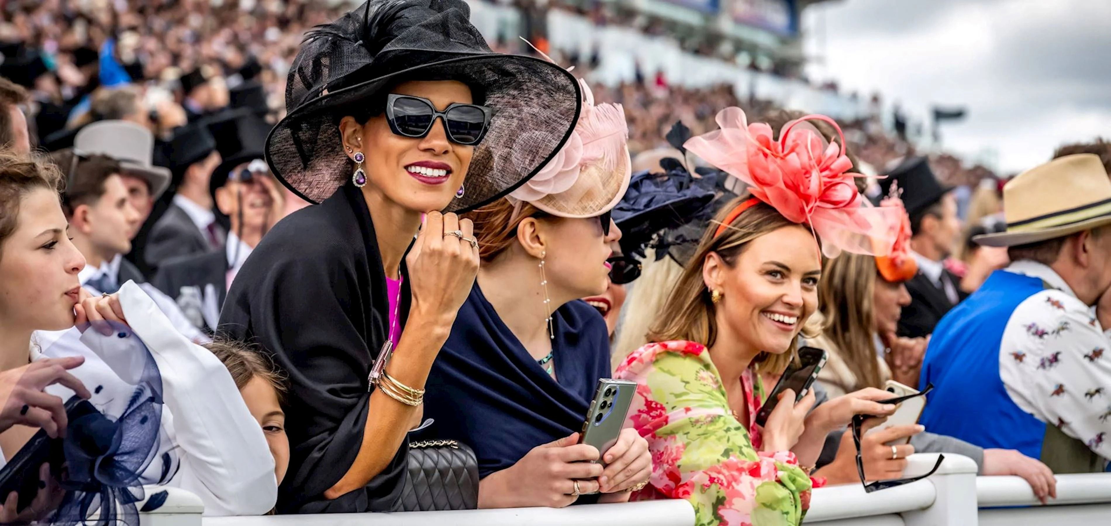 Racegoers along the rail at Epsom in wide-brimmed hats and fascinators, with men in morning dress and top hats behind them