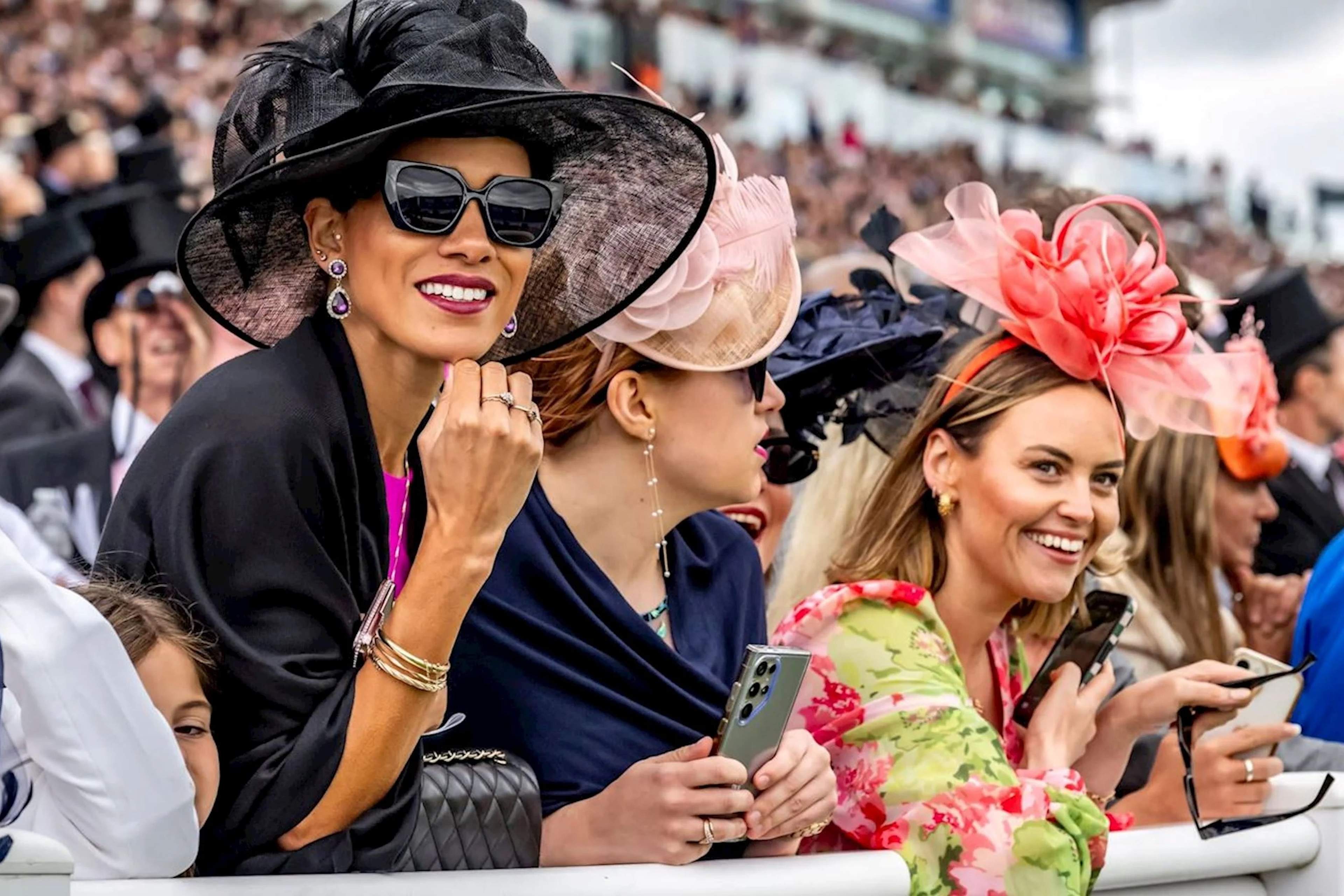 Racegoers along the rail at Epsom in wide-brimmed hats and fascinators, with men in morning dress and top hats behind them