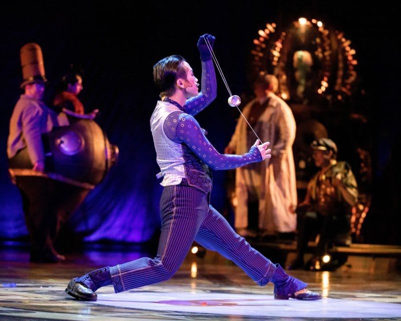 A KURIOS performer lunges across the stage with a yo-yo, the cast watching behind him