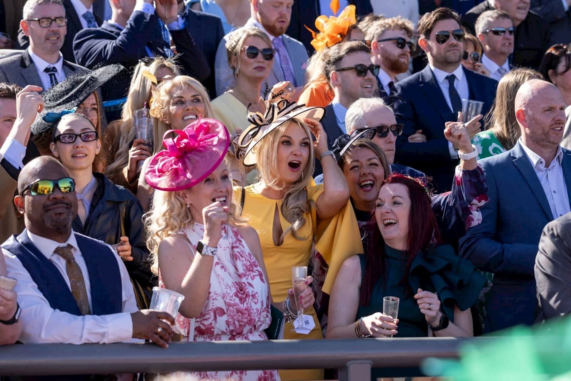 Racegoers at the Grand National cheering at the rail, the men in suits and the women in dresses with hats and fascinators