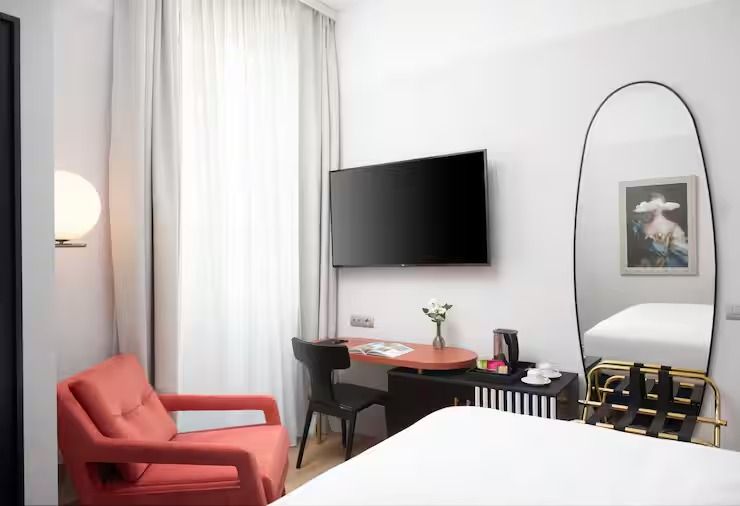 Guest room with a coral armchair by the window, a wall-mounted television above a coral-topped desk, and an arched mirror reflecting the bed