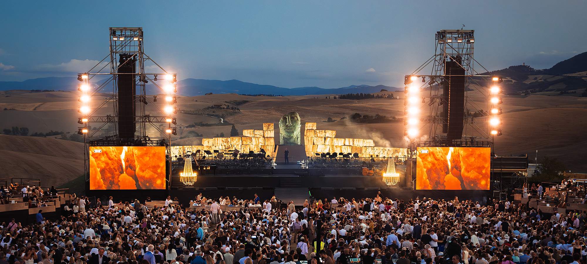 A singer and orchestra on a lit open-air stage at dusk, a large carved stone head behind them, a seated crowd in front and the Tuscan hills beyond