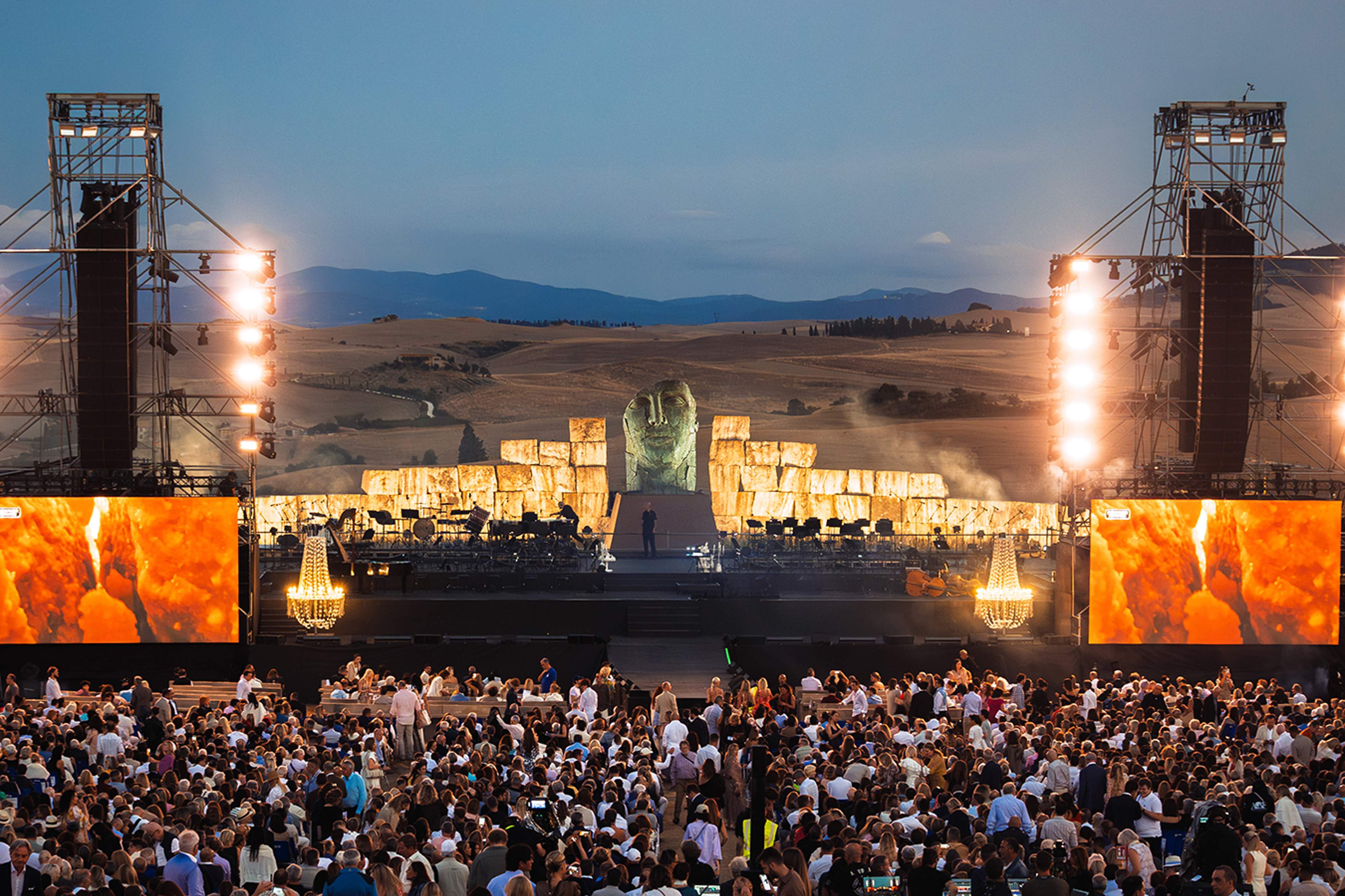 A singer and orchestra on a lit open-air stage at dusk, a large carved stone head behind them, a seated crowd in front and the Tuscan hills beyond