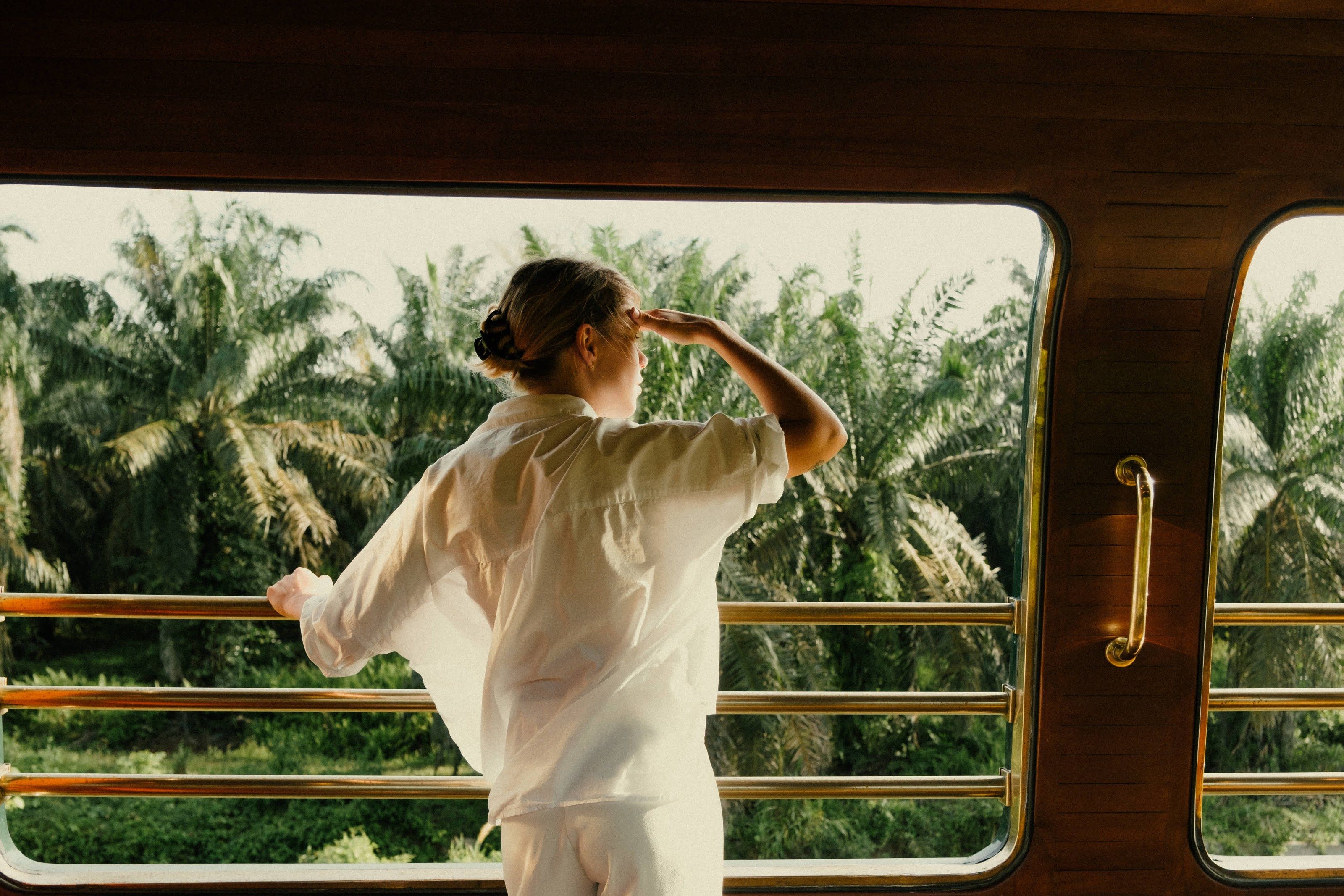 A guest shading her eyes at the open rail of the observation car, with sunlit palms beyond.