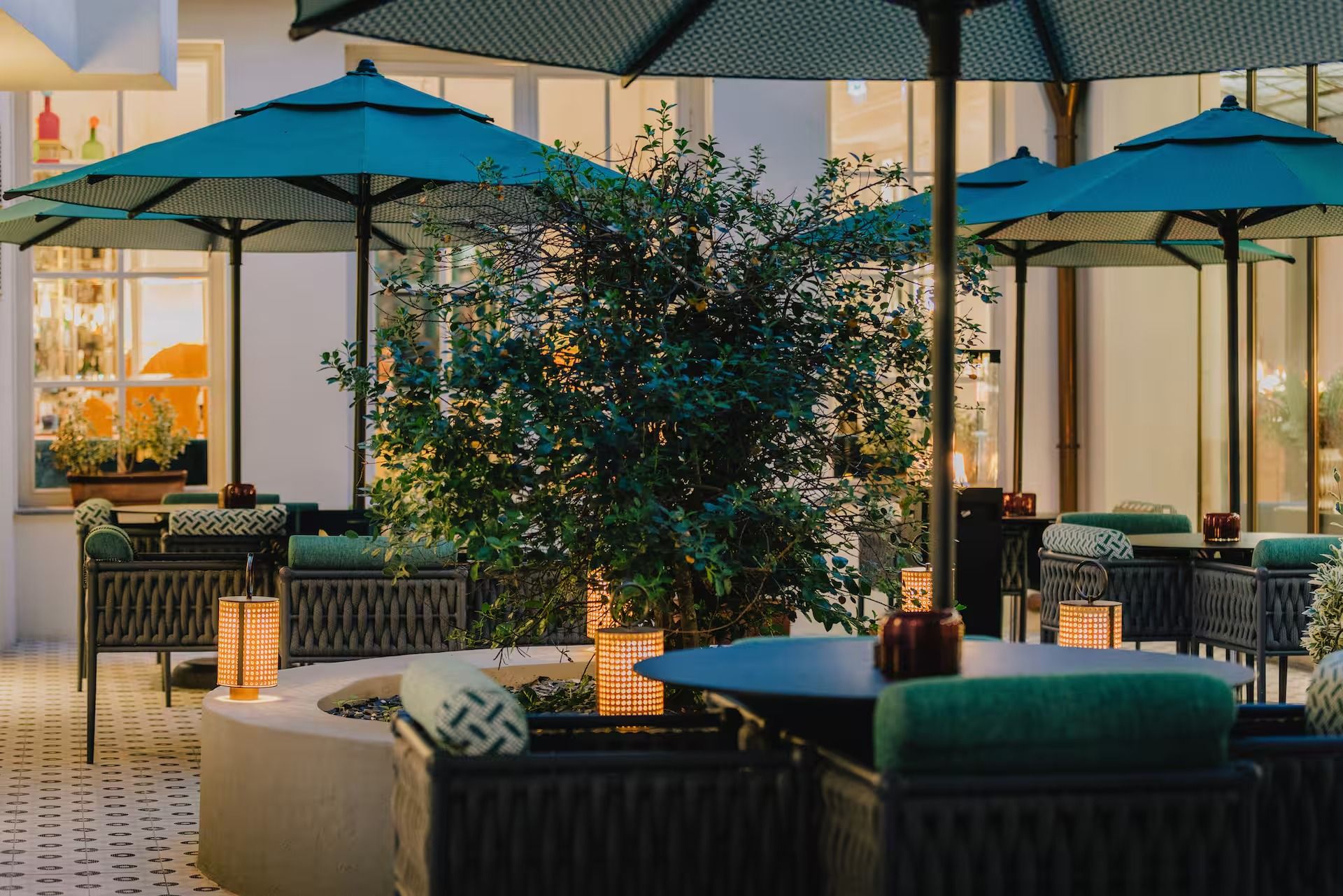 Courtyard terrace at dusk, with teal parasols, lit rattan lanterns on the tables and warm light from the bar windows