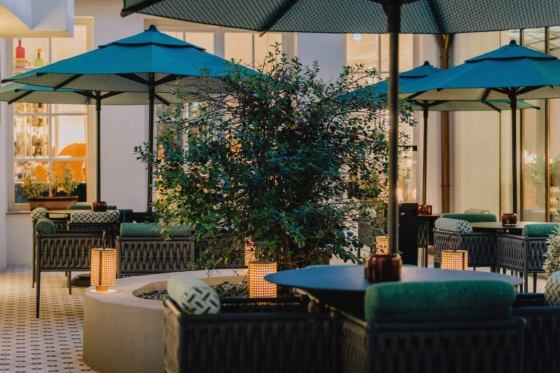 Courtyard terrace at dusk, with teal parasols, lit rattan lanterns on the tables and warm light from the bar windows