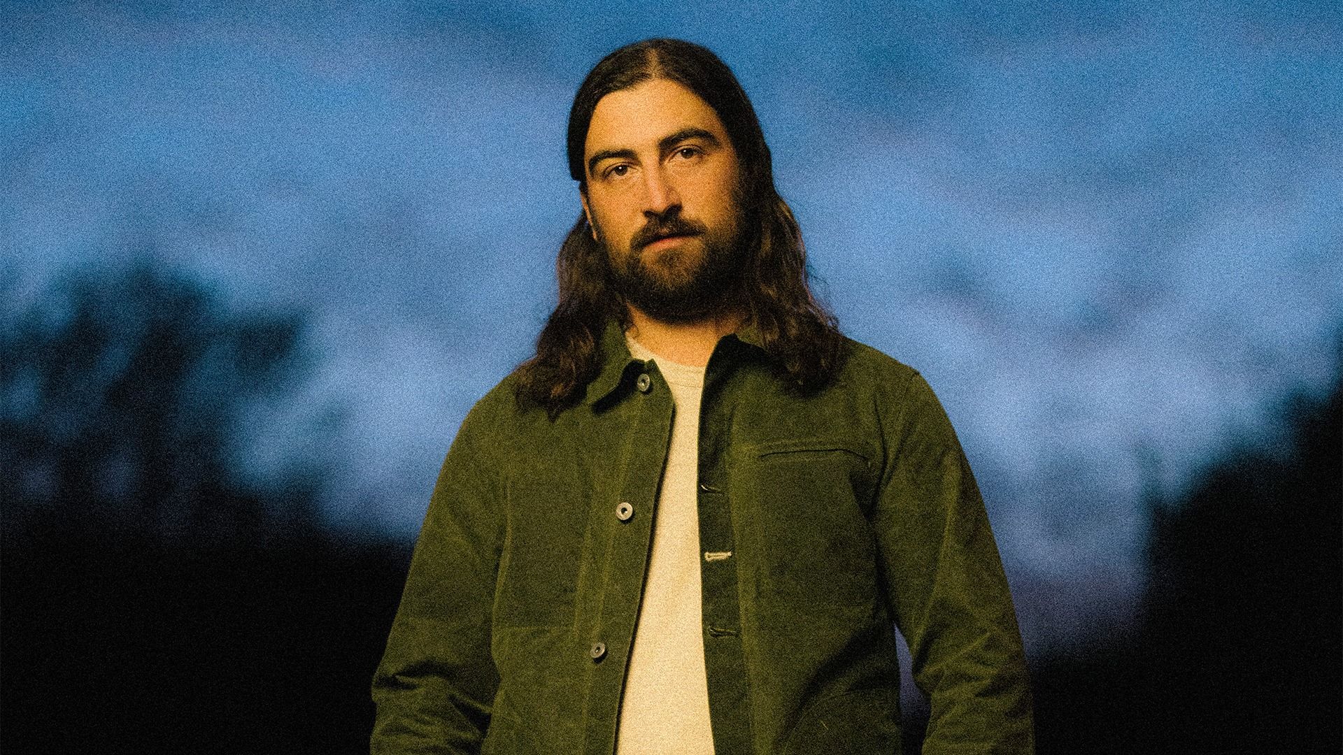 Noah Kahan in a green jacket, photographed outdoors at dusk