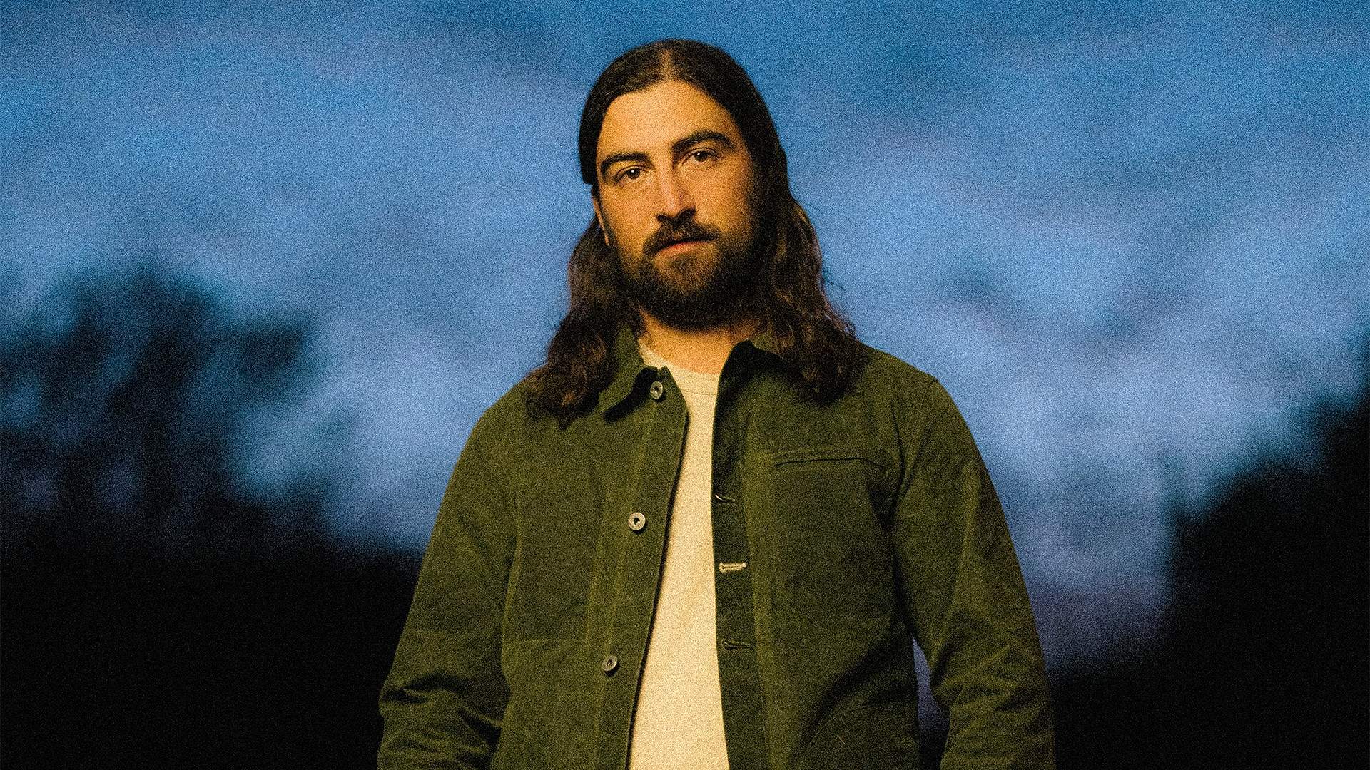 Noah Kahan in a green jacket, photographed outdoors at dusk