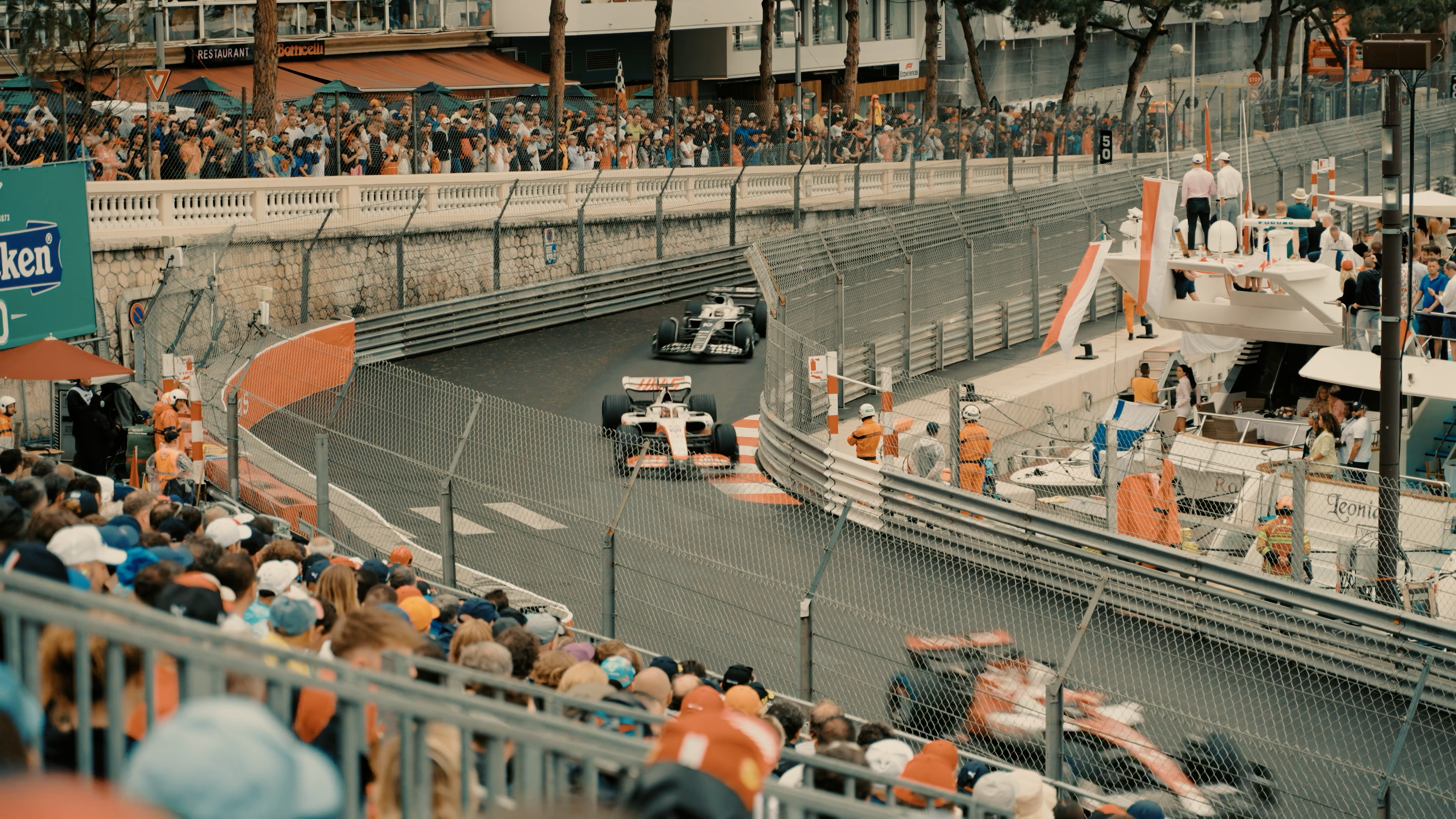 Two Formula 1 cars on track along the harbour front in Monaco, with spectators on a moored yacht beside the barrier