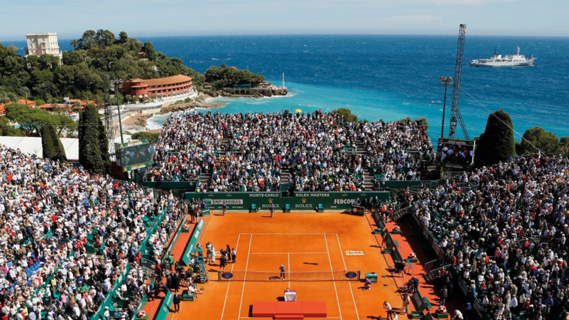 Court Rainier III full for a match at the Monte-Carlo Masters, with the Mediterranean and a headland behind the stands