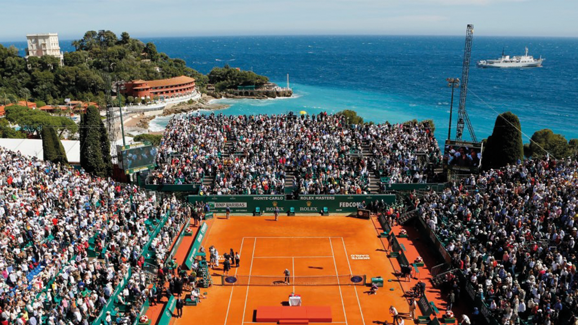 Court Rainier III full for a match at the Monte-Carlo Masters, with the Mediterranean and a headland behind the stands