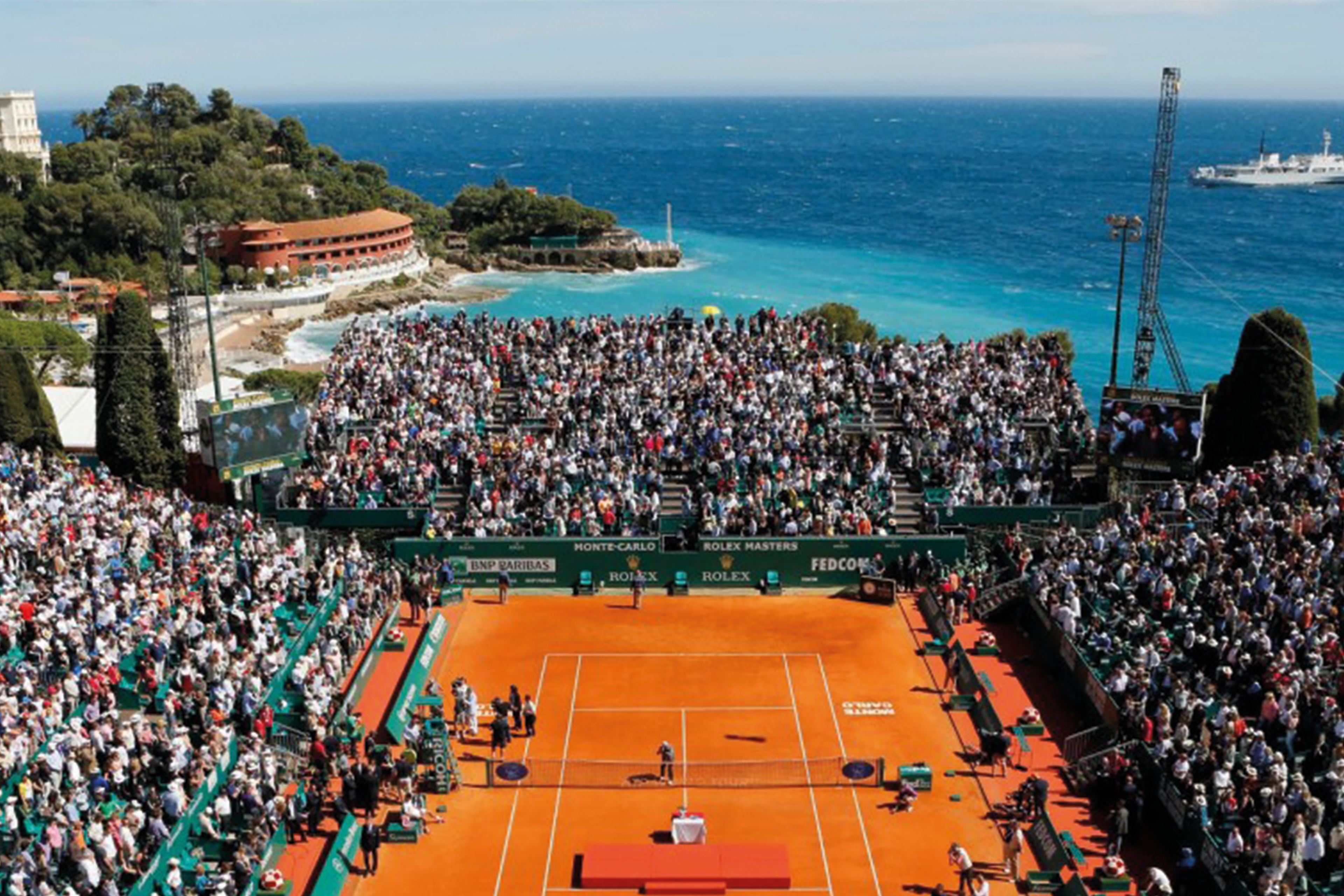 Court Rainier III full for a match at the Monte-Carlo Masters, with the Mediterranean and a headland behind the stands