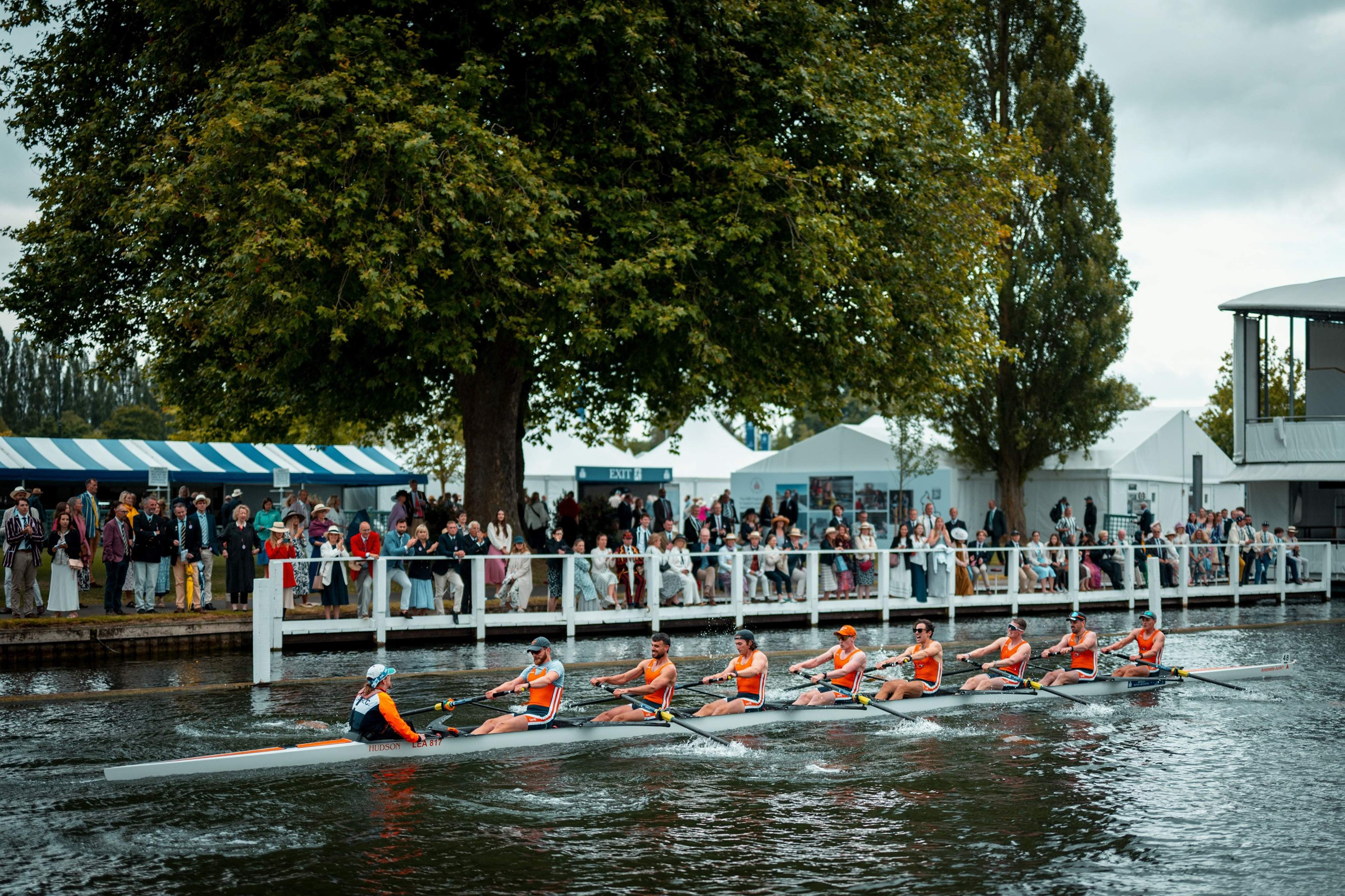 A rowing eight racing past the crowd on the bank at Henley, with marquees under the trees behind.