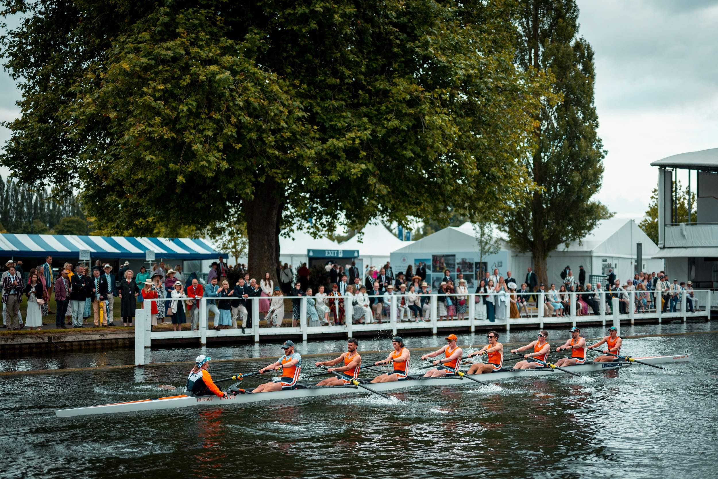 A rowing eight racing past the crowd on the bank at Henley, with marquees under the trees behind.