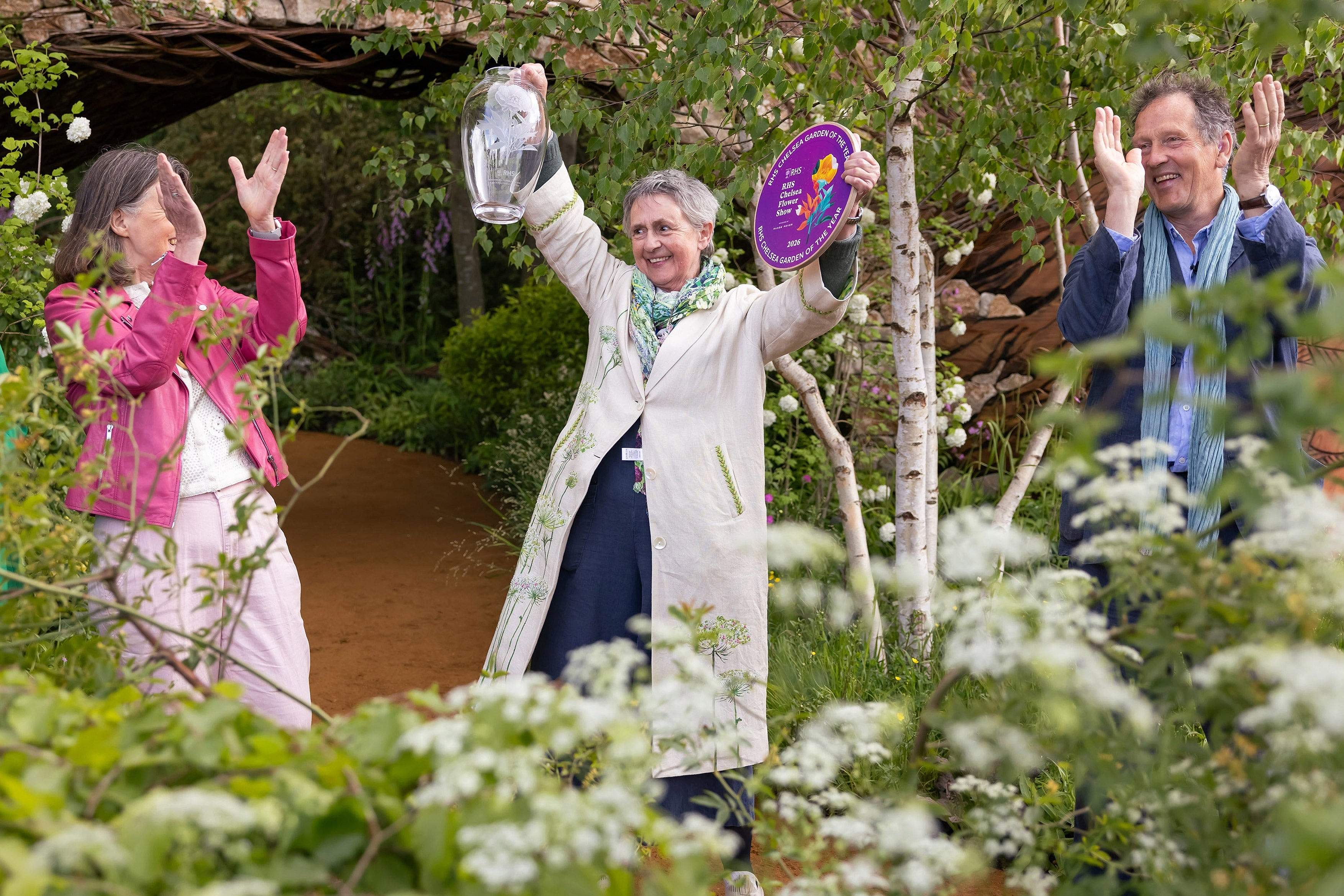 Three people raising their hands in a show garden at Chelsea, one in a pink jacket and two in light coats and scarves, among birch trees and cow parsley