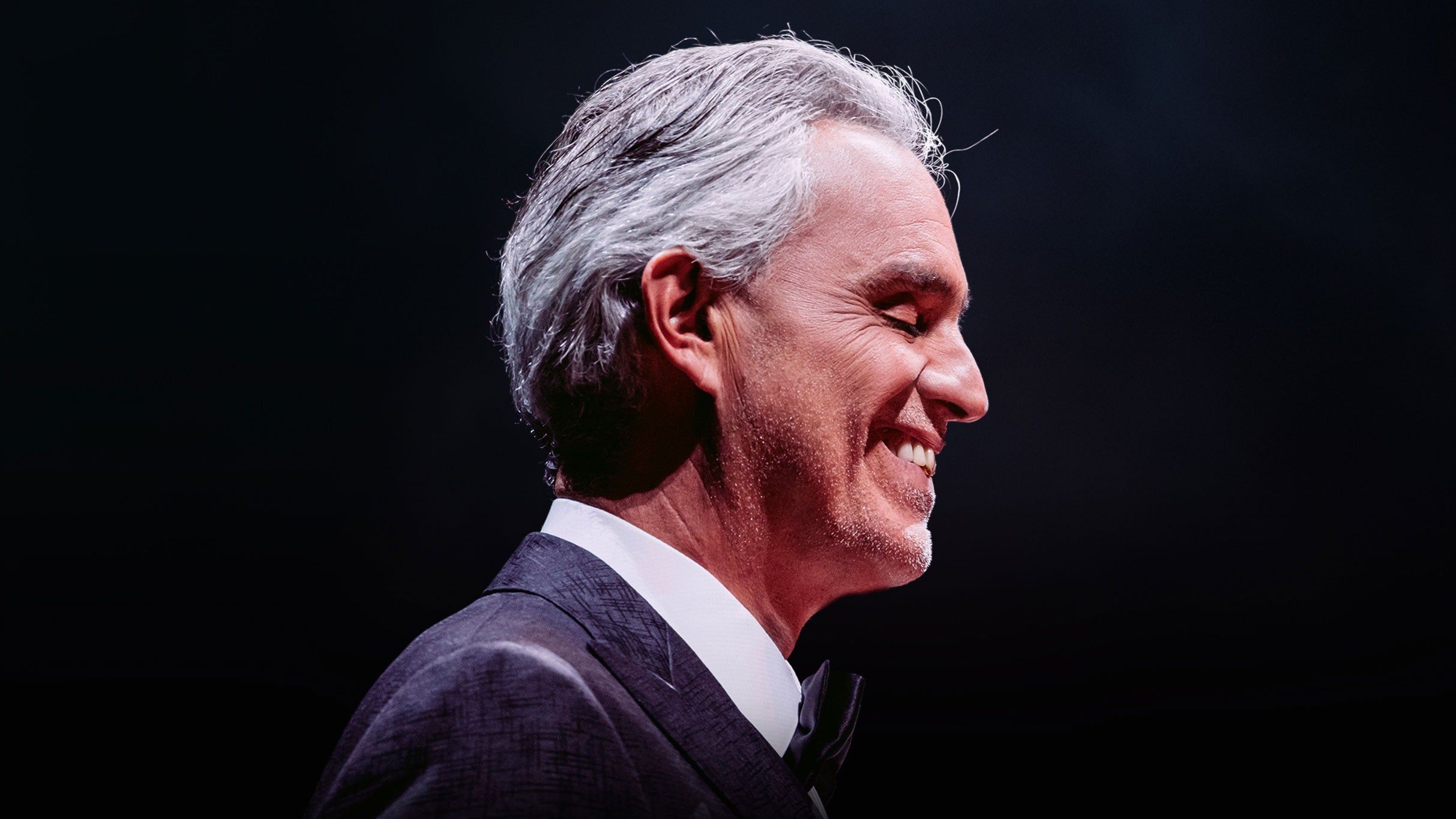 Andrea Bocelli in profile, smiling, in a dark suit and bow tie against a black stage backdrop