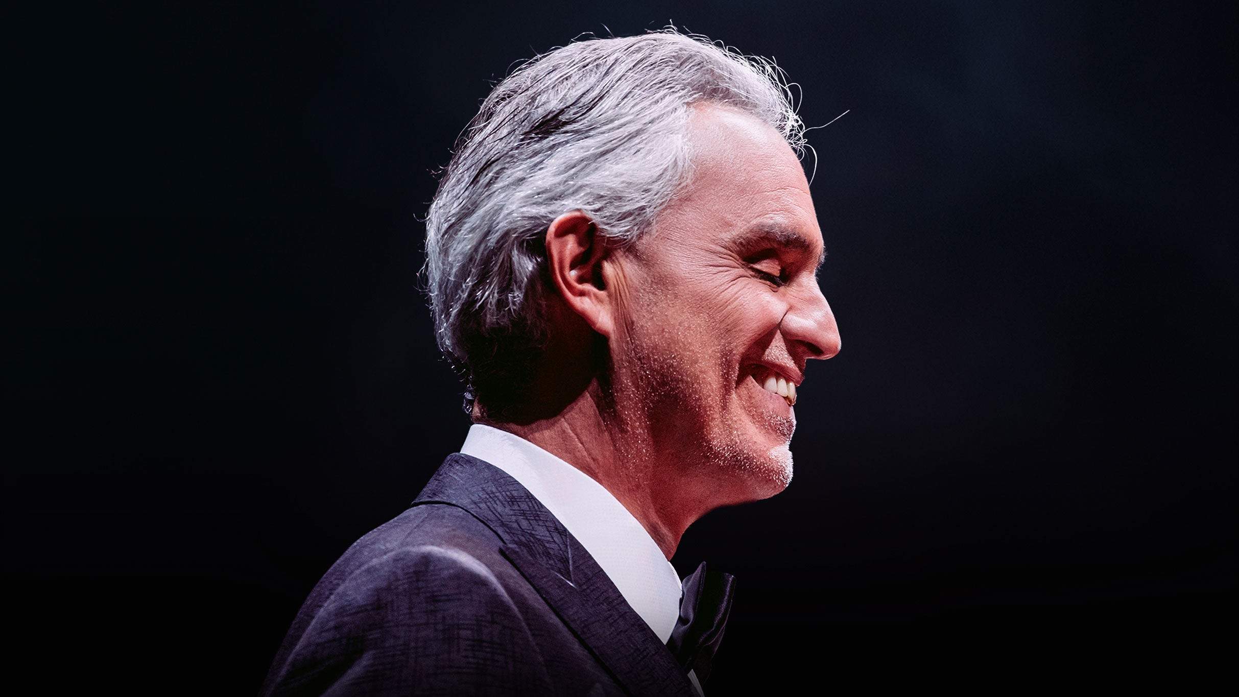 Andrea Bocelli in profile, smiling, in a dark suit and bow tie against a black stage backdrop
