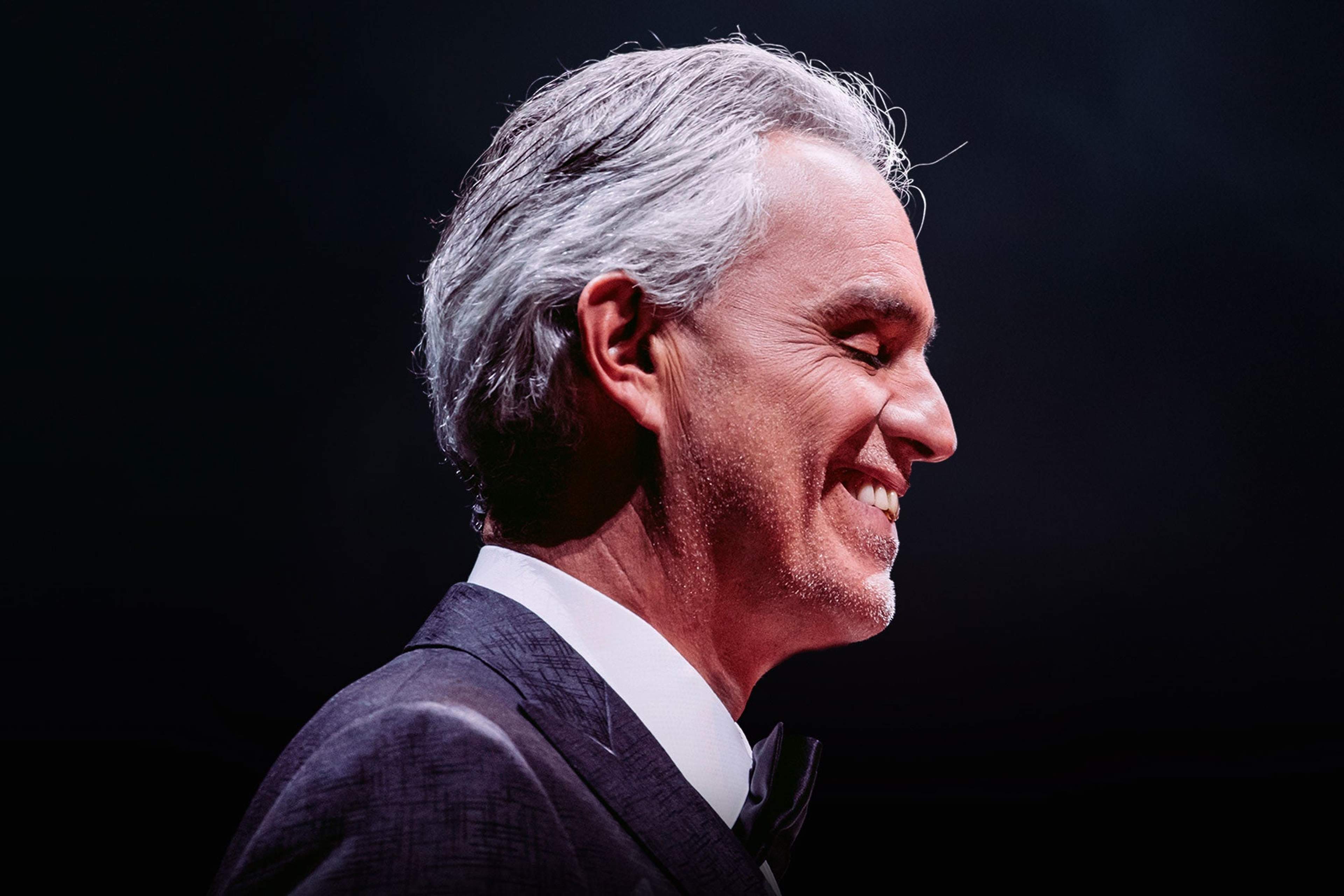 Andrea Bocelli in profile, smiling, in a dark suit and bow tie against a black stage backdrop