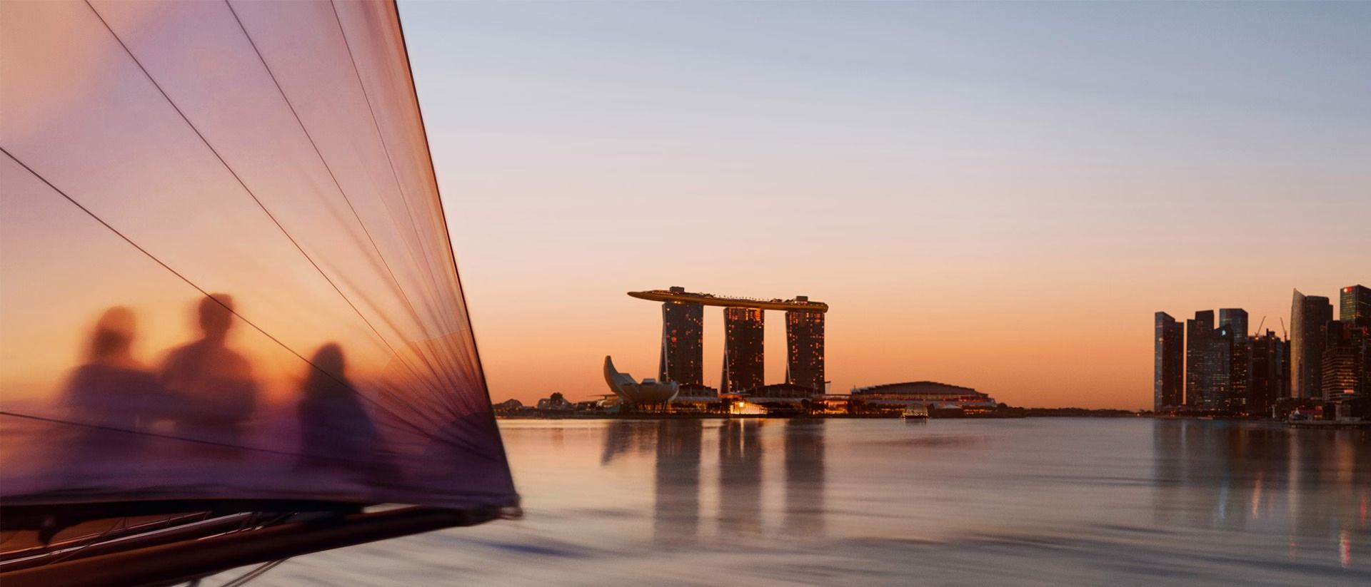 Silhouetted figures seen through a sail on Marina Bay at sunset, with Marina Bay Sands and the ArtScience Museum across the water.