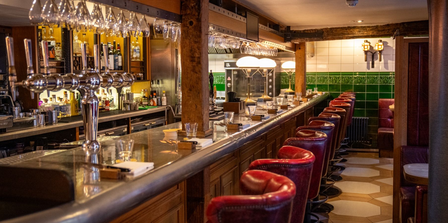 A pub bar with red leather stools, green tiles and an open kitchen behind the counter