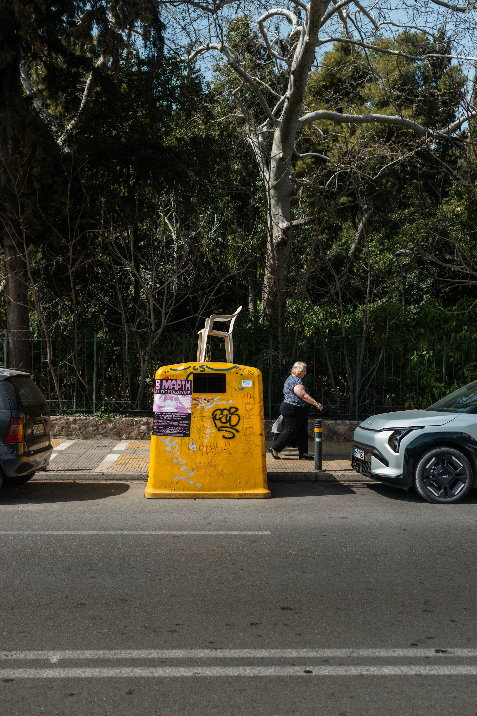 A woman walks past a yellow dumpster with a chair casually balanced on top, graffiti-covered walls behind her — Athens street life at its most honest