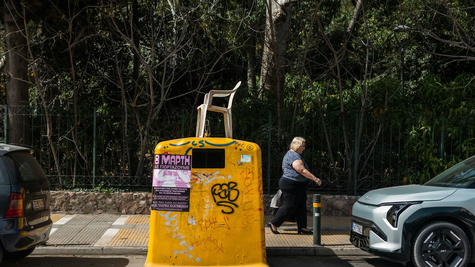 A woman walks past a yellow dumpster with a chair casually balanced on top, graffiti-covered walls behind her — Athens street life at its most honest