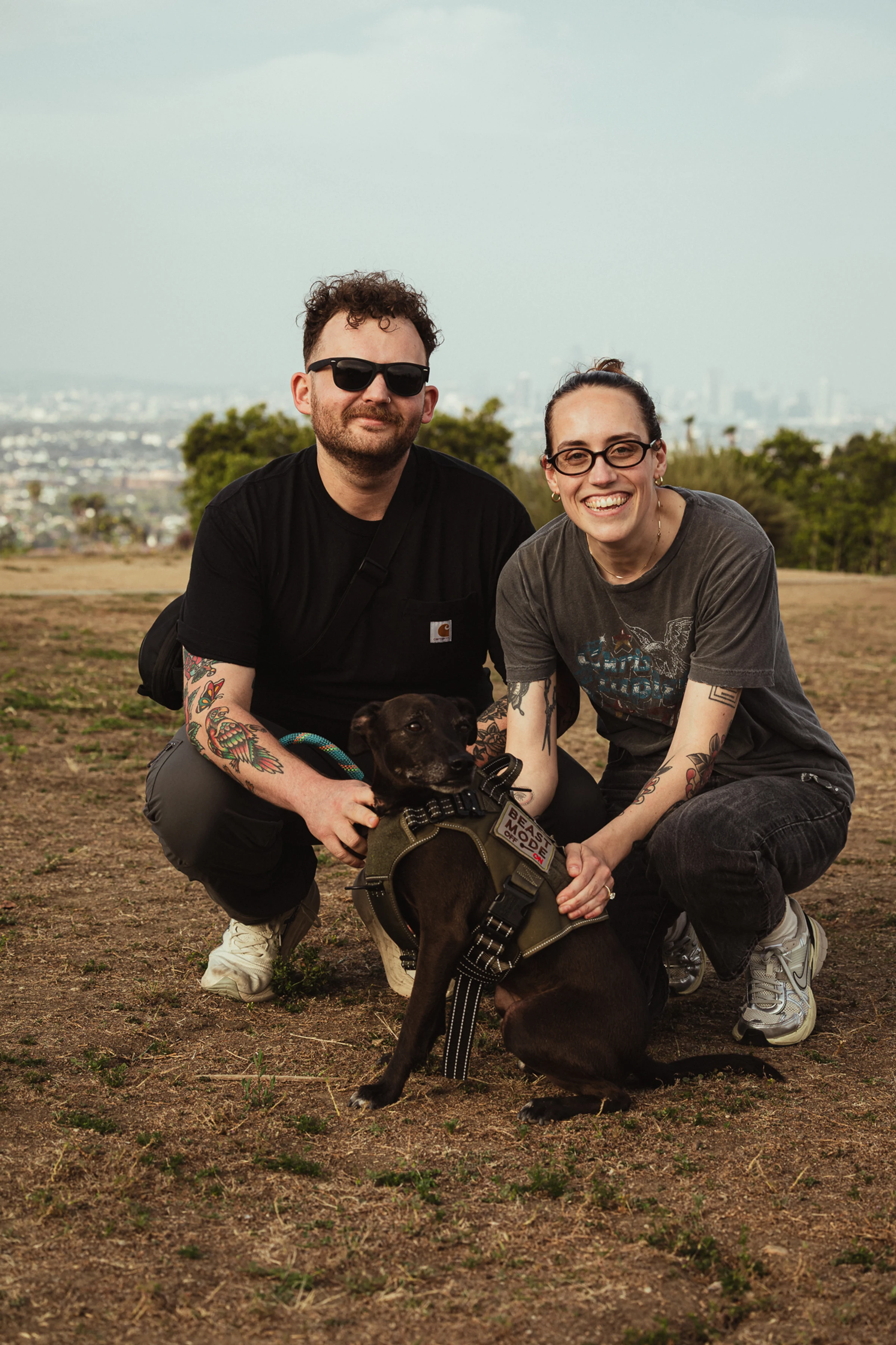 A smiling couple crouches on a grassy hill with a black dog in a harness. The man wears sunglasses; the city skyline is faintly visible in the distance.