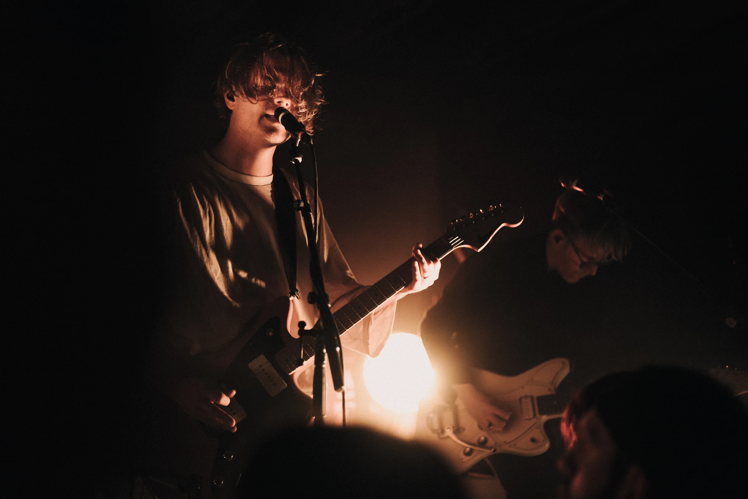 Two musicians perform on stage under moody lighting. The lead singer plays guitar and sings into a microphone. A warm, atmospheric glow surrounds them.