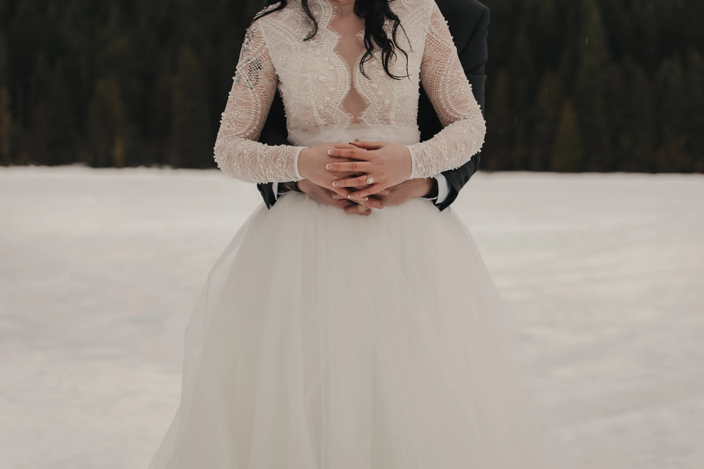 A bride in a lace wedding dress with long, wavy hair stands on snow, embraced by a groom in winter. The mood is intimate and serene, with a forest backdrop.